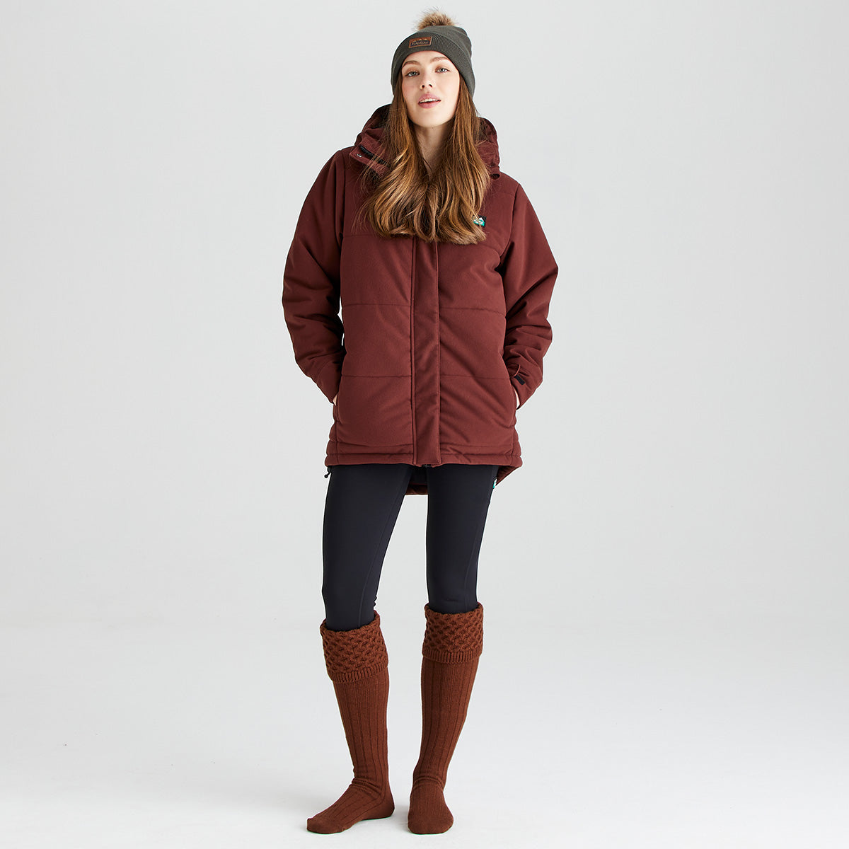 Lifestyle image of a person wearing rust-brown welly socks with long ribbed legs and chunky cable-knit turnover cuffs. The socks are paired with black leggings, a burgundy insulated outdoor jacket, and a dark knit beanie. Photographed against a light grey studio background, the image highlights the welly socks' knee-high fit, textured knit construction, and classic country styling, designed to provide warmth and comfort when worn with wellington boots in colder conditions.

