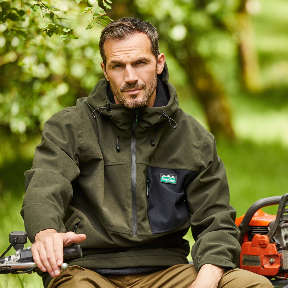 Man wearing a green waterproof smock with black chest patch Ridgeline Clothing Logo. Sitting on quad bike with trees in the background