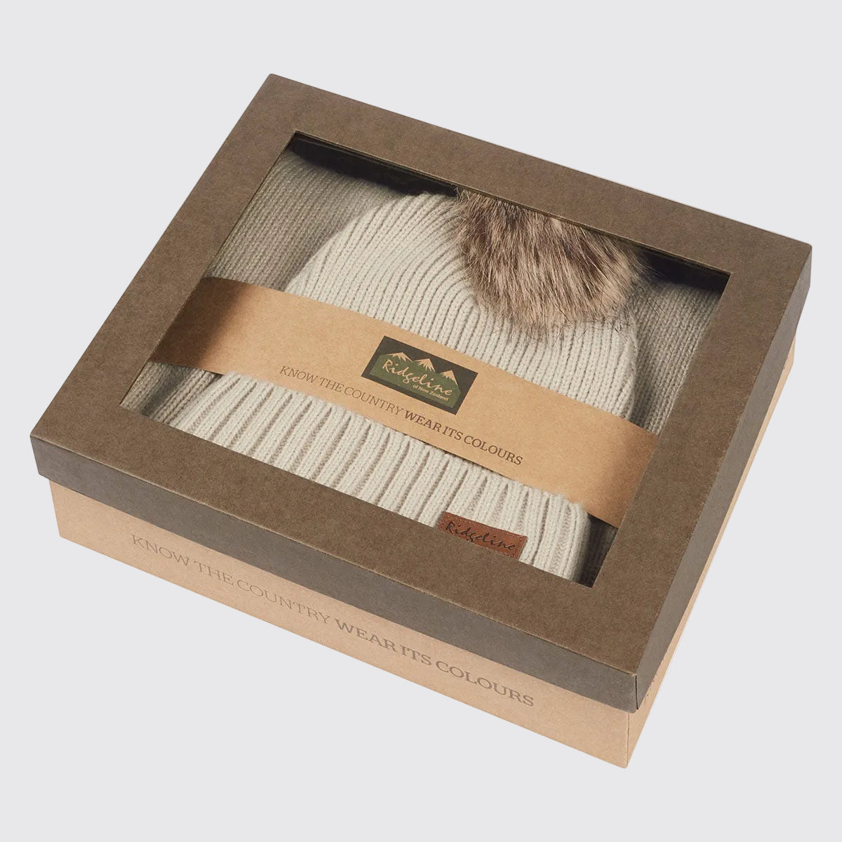 Cream ribbed knit beanie hat with faux fur pom pom in windowed gift box, angled view.