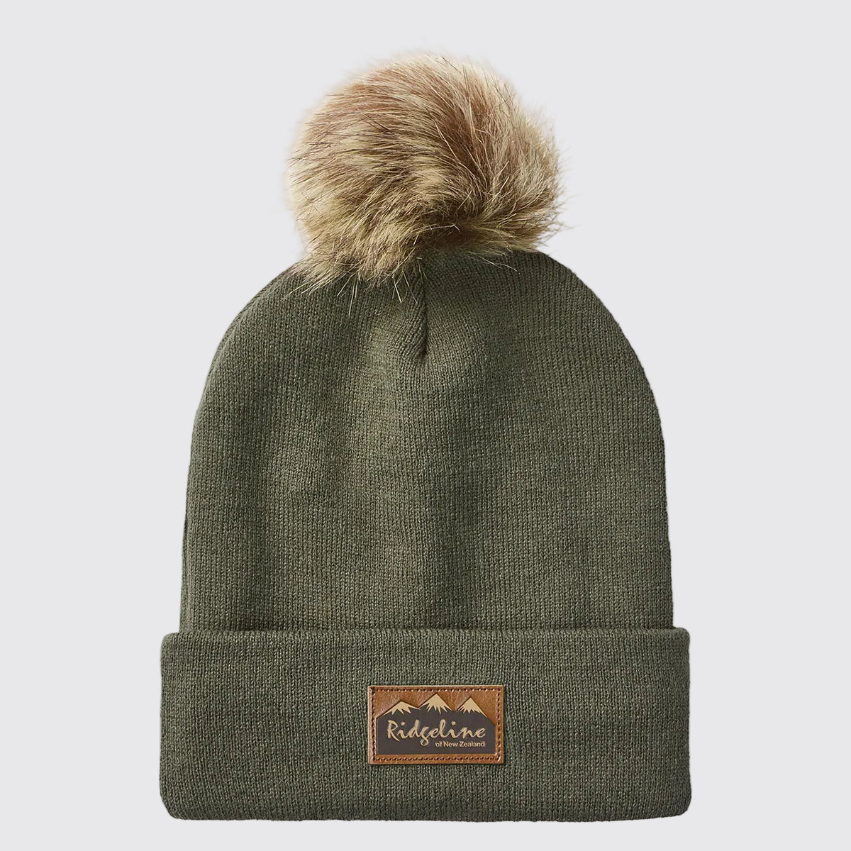 Front view of a greenish-brown ribbed knit beanie with a folded cuff and a large faux-fur pom-pom attached at the crown, displayed against a light grey background. A brown faux-leather Ridgeline of New Zealand logo patch with a mountain graphic is centered on the cuff. The fine-knit texture, soft pom-pom detail, and classic cuffed design are clearly visible, giving the beanie a warm, stylish look suitable for cold-weather outdoor wear.
