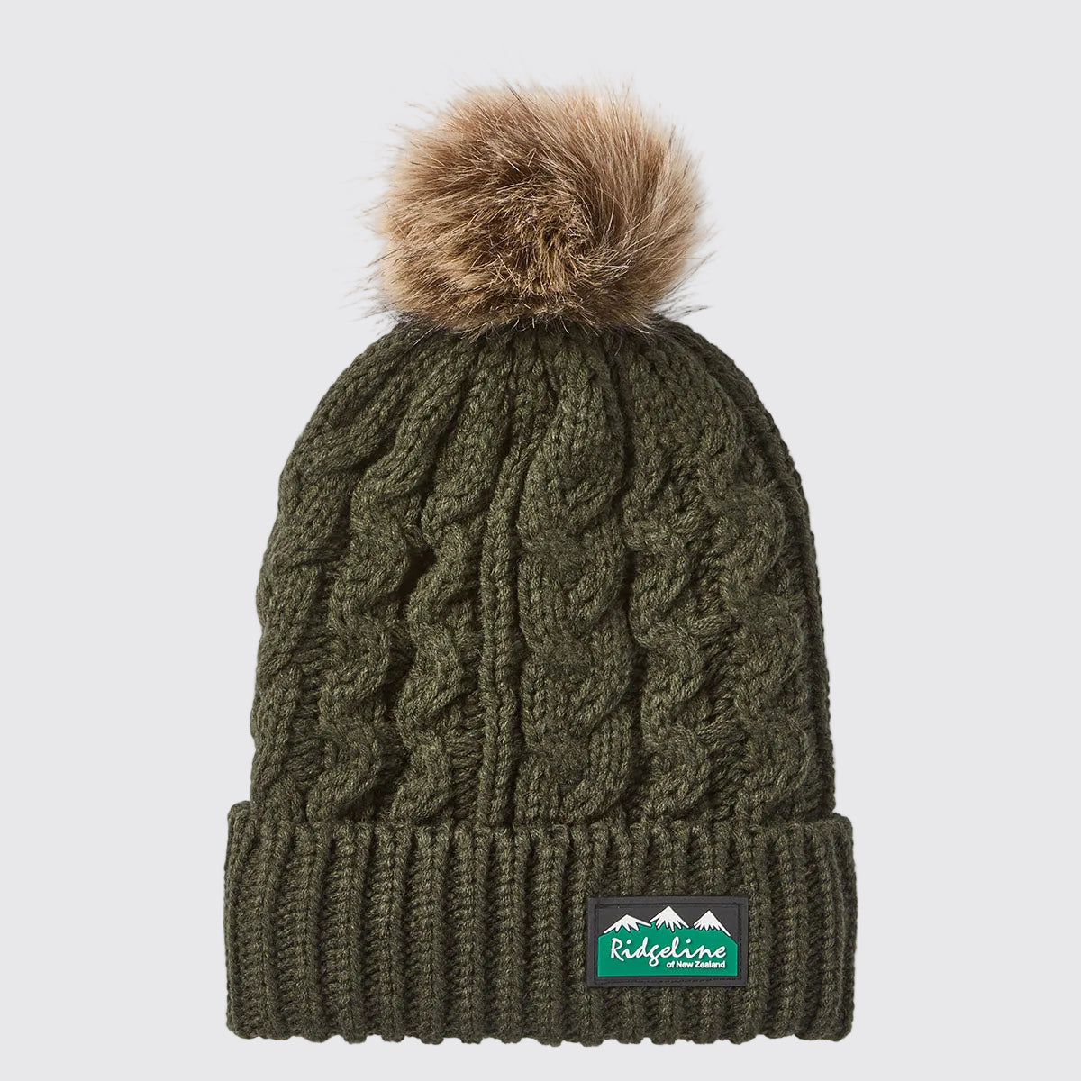 Front view of an olive green cable-knit beanie with a ribbed cuff and a large faux-fur pom-pom attached at the crown, displayed against a light grey background. A Ridgeline of New Zealand logo patch is positioned on the front cuff. The beanie features a textured cable-knit pattern throughout, adding depth and visual interest, while the soft pom-pom provides a stylish cold-weather detail. The thick knit construction and cuffed design highlight warmth, comfort, and outdoor-ready practicality.
