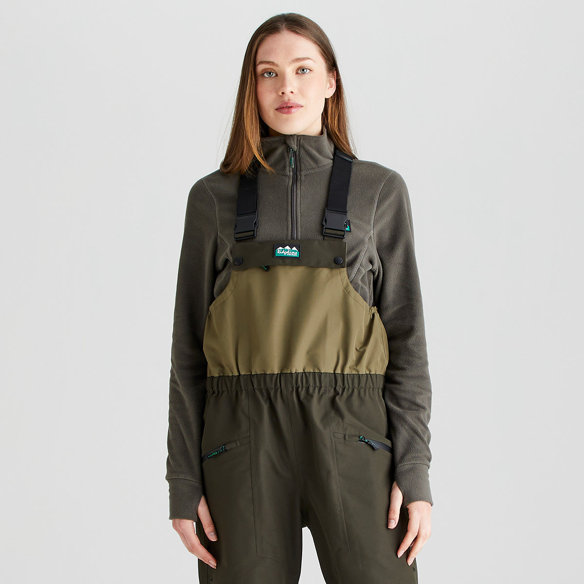 Front-facing studio image of a person wearing olive-green chest waders with adjustable black shoulder straps over a dark olive zip-neck fleece top. The waders feature a two-tone design with a lighter olive upper section, elasticized waist, and zippered pockets on the front. A small logo patch is visible on the chest panel. The outfit is shown against a light gray background with even lighting, highlighting the fit, fabric, and functional details of the outdoor apparel.
