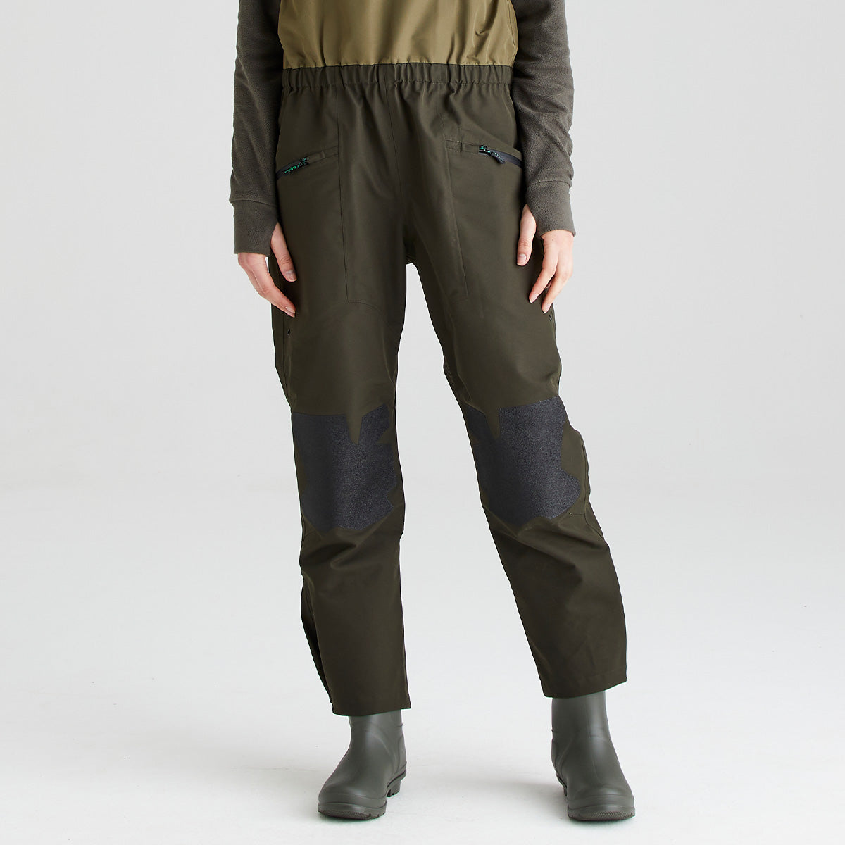 Lower-body view of olive-green waterproof bib and brace trousers with integrated rubber boots, shown against a light gray studio background. The bib and brace features an elasticated waist, angled zippered hand pockets, and reinforced knee panels in a contrasting dark textured material for enhanced durability and protection. The image highlights the fit, rugged construction, a