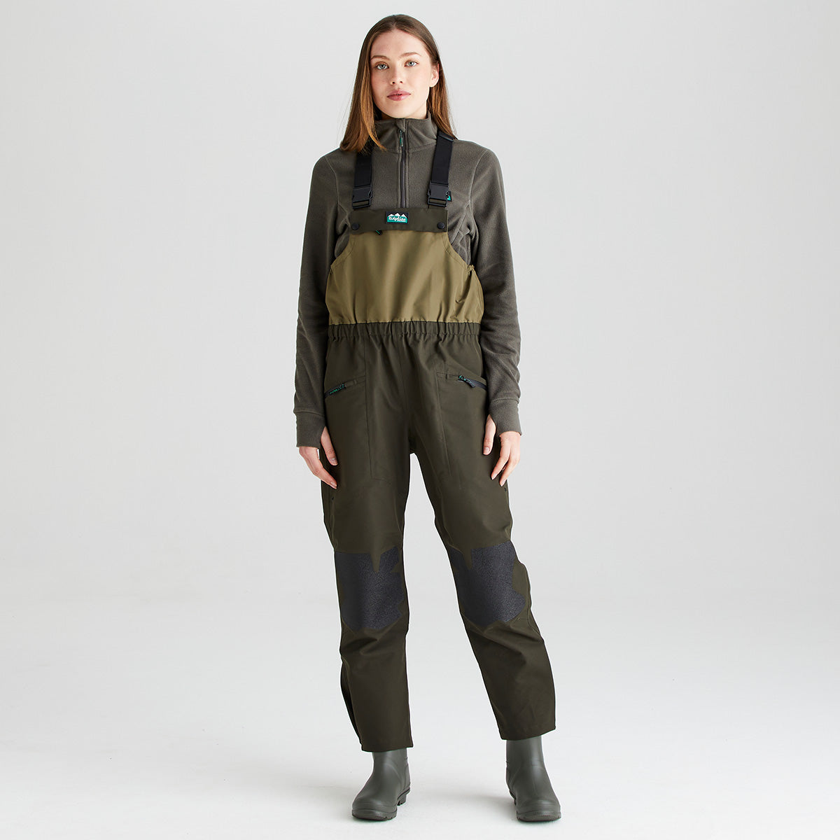 Full-length front view of an olive-green waterproof bib and brace with integrated rubber boots, shown against a light gray studio background. The garment features adjustable black shoulder straps, a two-tone olive bib panel, an elasticated waist, angled zippered pockets, and reinforced knee panels in a contrasting dark textured material for enhanced durability. The image highlights the overall fit, protective construction, and practical outdoor performance features of the bib and brace.
