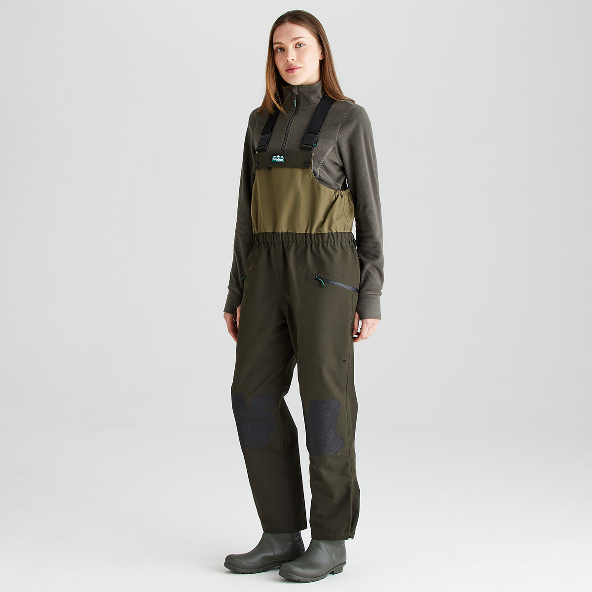 Full-length studio image of a person wearing olive-green waterproof bib and brace trousers with integrated rubber boots and adjustable black shoulder straps. The bib and brace features a two-tone olive design, elasticated waist, zippered chest and hand pockets, and reinforced knee panels for added durability. Shown against a light gray background, the image provides a clear front three-quarter view of the garment's fit, construction, and practical outdoor performance features.
