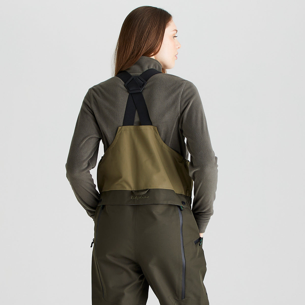 Rear three-quarter view of an olive-green waterproof bib and brace worn over a dark olive fleece top, shown against a light gray studio background. The bib and brace features adjustable black shoulder straps with a crossover back design, a two-tone olive construction, and durable waterproof fabric. The image highlights the rear bib panel, strap configuration, and overall fit across the back and upper body.
