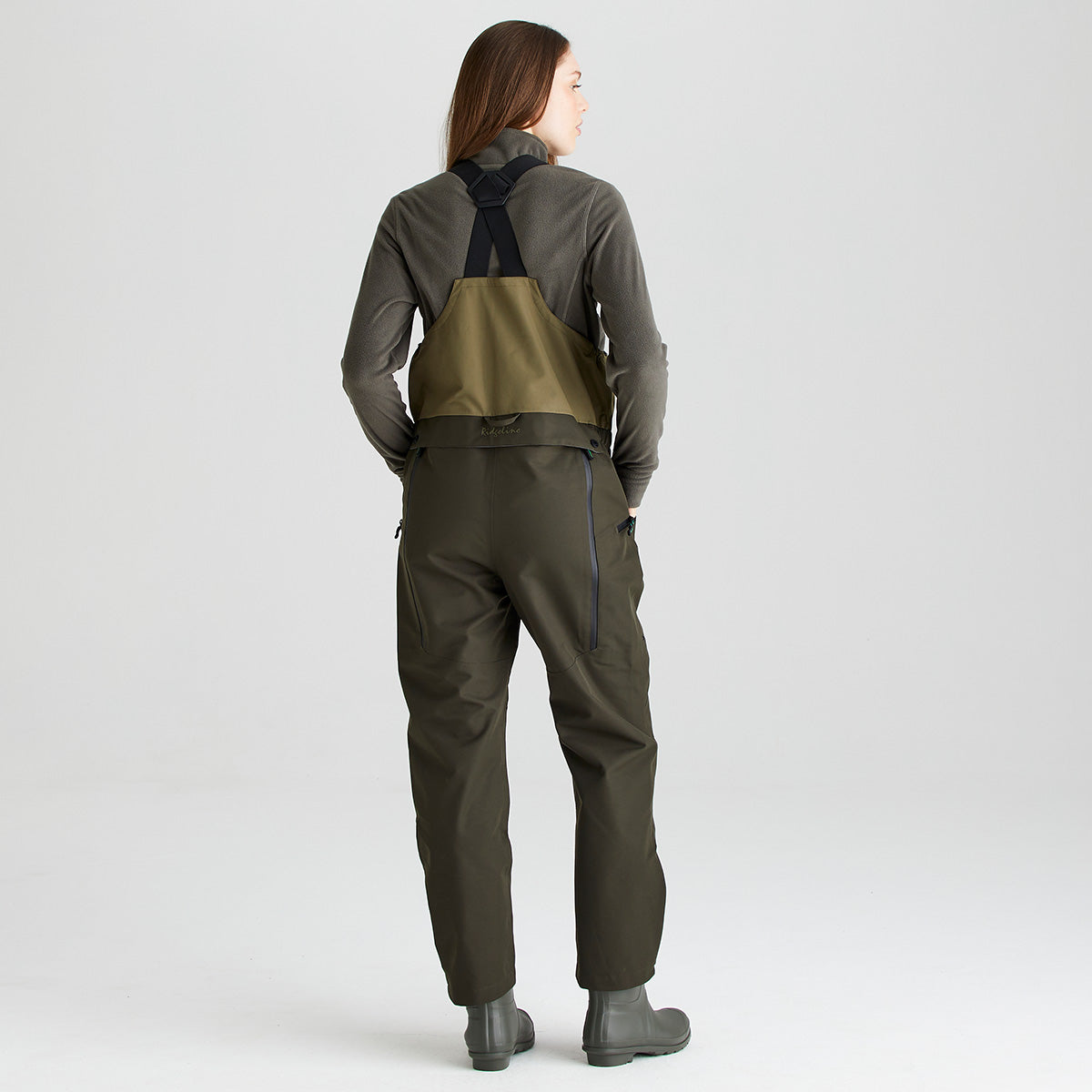 Full-length rear view of an olive-green waterproof bib and brace with integrated rubber boots, shown against a light gray studio background. The bib and brace features a two-tone olive design, adjustable black shoulder straps with a crossover back arrangement, and durable waterproof construction. Rear zippered pockets are visible on either side, while the straight-leg fit and integrated boots provide practical protection for outdoor activities. 