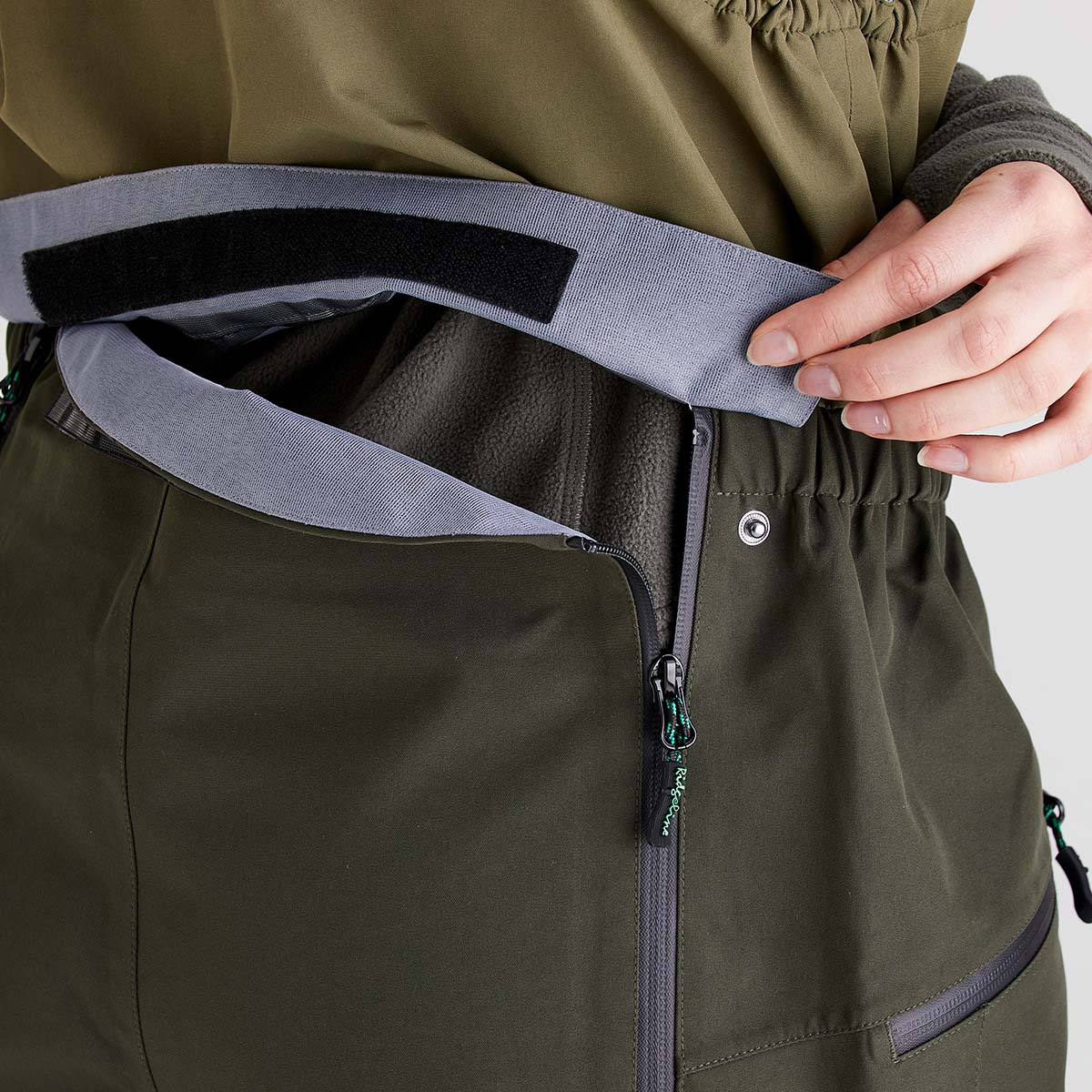 Close-up detail of an relief system integrated into olive-green chest waders. A hand is lifting a gray protective flap with a hook-and-loop fastening to reveal the emergency relief opening beneath. The design includes a vertical waterproof zipper, elasticized waist section, and durable performance fabric. The image focuses on the accessibility and functionality of the relief system, highlighting the protective flap and zippered opening against a light studio background.
