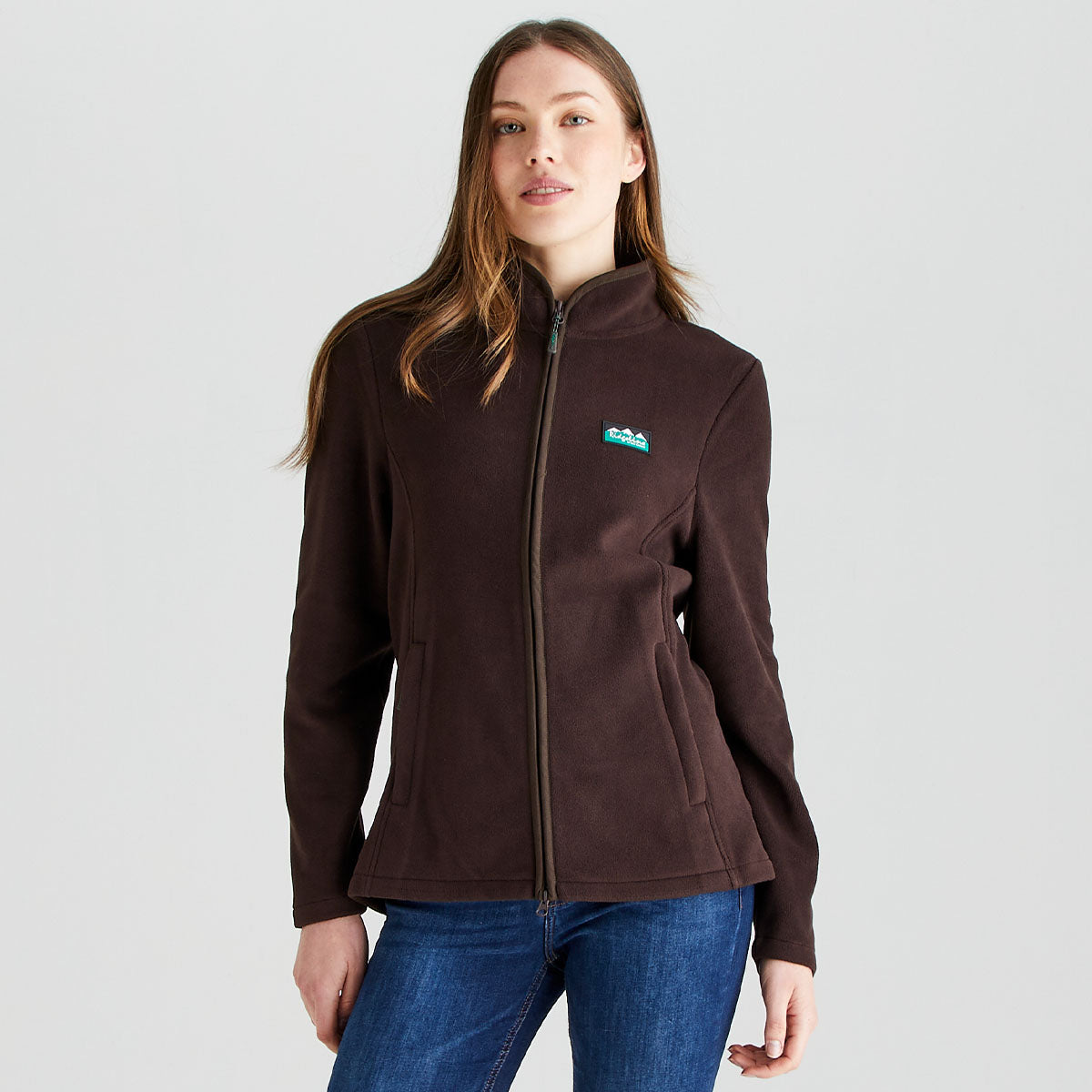 Front view of a dark brown fleece jacket worn with blue jeans. The jacket features a high stand-up collar, a full-length front zip fastening, and two discreet zipped hand pockets set into the front seams. A small Ridgeline logo patch is positioned on the left chest. The fitted silhouette and soft fleece fabric create a streamlined, comfortable look suitable for outdoor and casual wear. Photographed against a plain light-grey studio background, the image highlights the jacket’s simple design 
