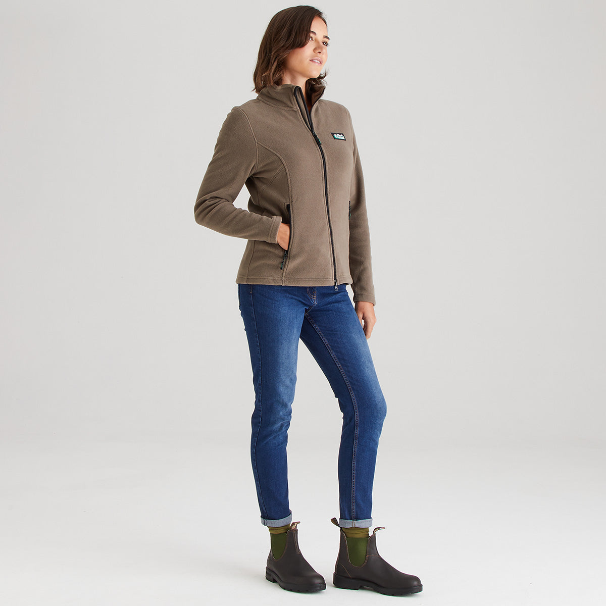 Woman wearing a brown jacket, blue jeans, and black boots on a light gray background