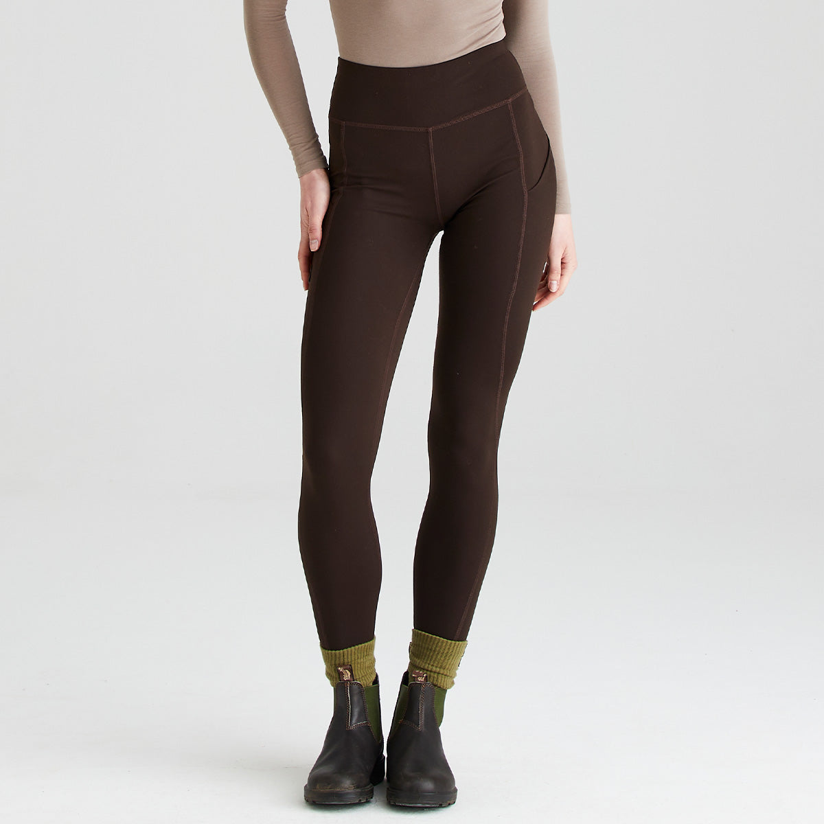 Front view of dark brown performance leggings worn with black ankle boots and olive-green socks. The leggings feature a high-rise, wide waistband for a supportive fit and contoured seam detailing along the legs. A side pocket is visible on the upper thigh, adding practical storage. The image is photographed in a studio against a plain light-grey background, highlighting the streamlined, close-fitting design.