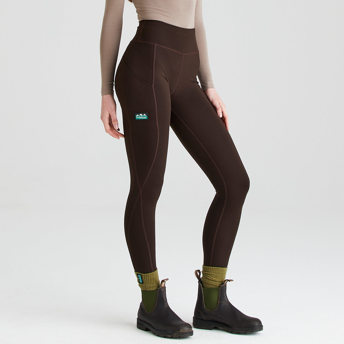 Three-quarter view of dark brown high-waisted leggings worn with black ankle boots and olive-green socks. The leggings feature a wide supportive waistband, body-contouring seam detailing, and a side thigh pocket for convenient storage. A small branded logo is positioned on the upper thigh. Photographed against a plain light-grey studio background, the image highlights the leggings' streamlined fit, stretch fabric, and practical activewear design.
