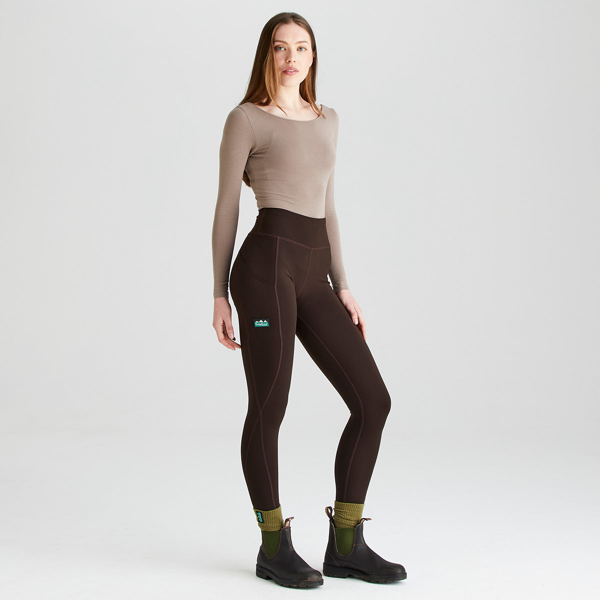 Full-length studio image of a person wearing dark brown high-waisted leggings with a fitted beige long-sleeve top, olive-green socks, and black ankle boots. The leggings feature a wide supportive waistband, curved seam detailing, and side thigh pockets for convenient storage. Shown in a three-quarter pose against a plain light-grey background, the image highlights the leggings' streamlined fit, stretch fabric, and practical activewear design.