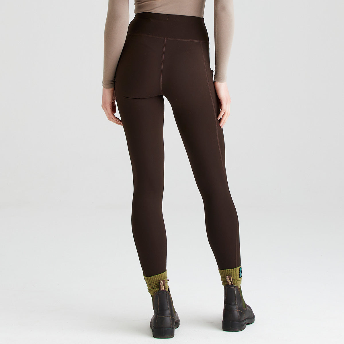 Rear view of dark brown high-waisted leggings worn with black ankle boots and olive-green socks. The leggings feature a wide supportive waistband, a smooth close-fitting silhouette, and side thigh pockets visible from the back. Minimal seam detailing enhances the streamlined design and flexible fit. The image is photographed against a plain light-grey studio background, highlighting the leggings' shape, coverage, and practical activewear styling.