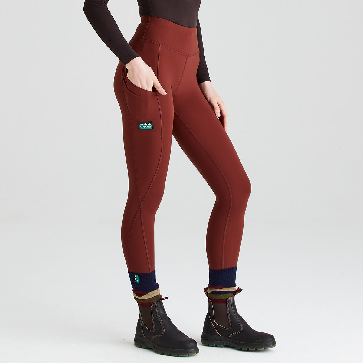 Three-quarter view of rust-red performance leggings worn with black ankle boots and layered navy and striped socks. The leggings feature a high-rise, wide waistband, curved contour seams, and a large side thigh pocket, shown with a hand resting inside the pocket to demonstrate functionality. A small branded logo patch is positioned on the upper thigh. Photographed against a plain light-grey studio background, the image highlights the leggings’ supportive fit, stretch fabric, and practical storage design.