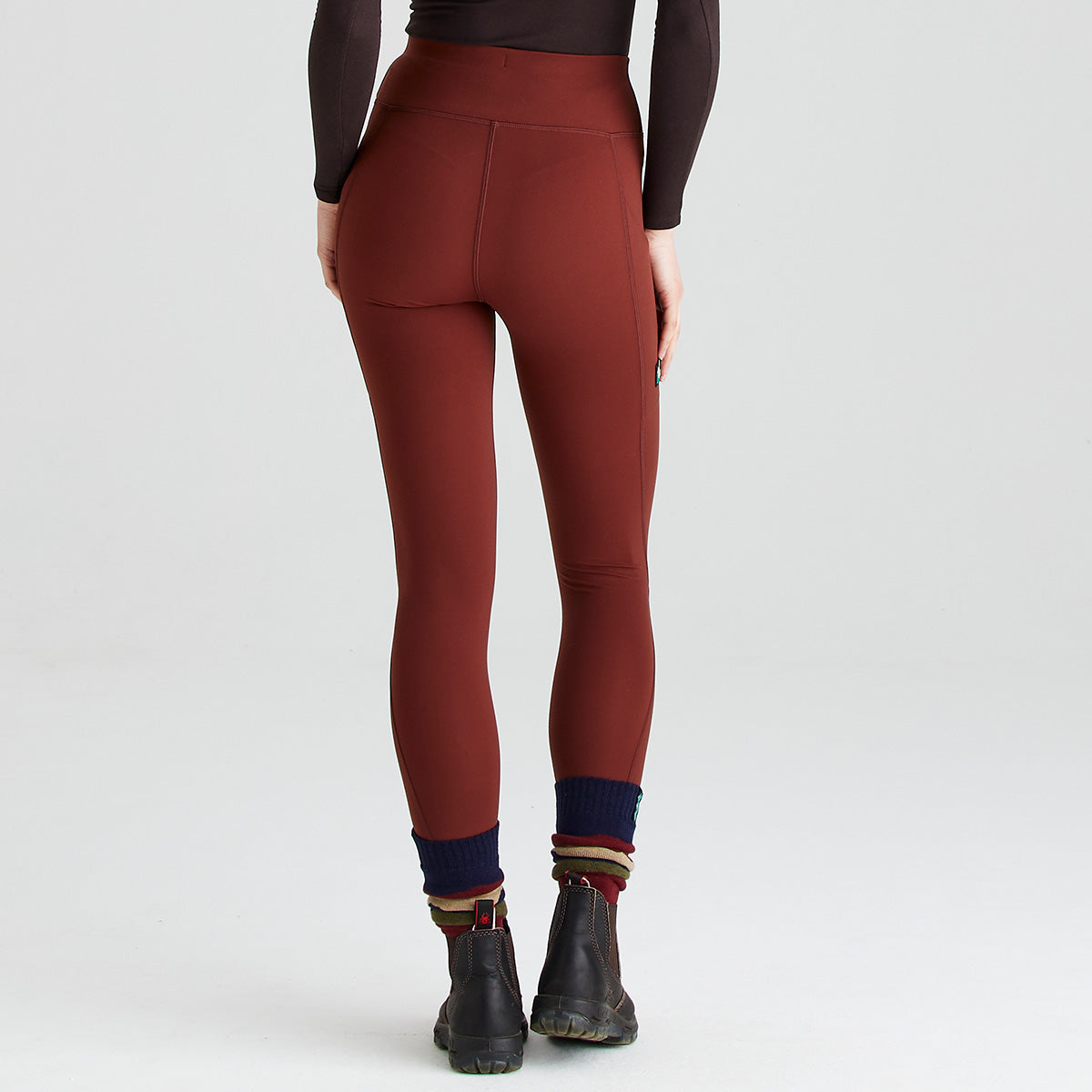 Rear view of rust-red high-waisted performance leggings worn with black ankle boots and navy socks with striped cuffs. The leggings feature a wide supportive waistband, a smooth close-fitting silhouette, and side thigh pockets visible along the outer legs. Minimal contour seam detailing enhances the streamlined design and flexible fit. Photographed against a plain light-grey studio background, the image highlights the leggings’ shape, coverage, and practical activewear styling from the back.