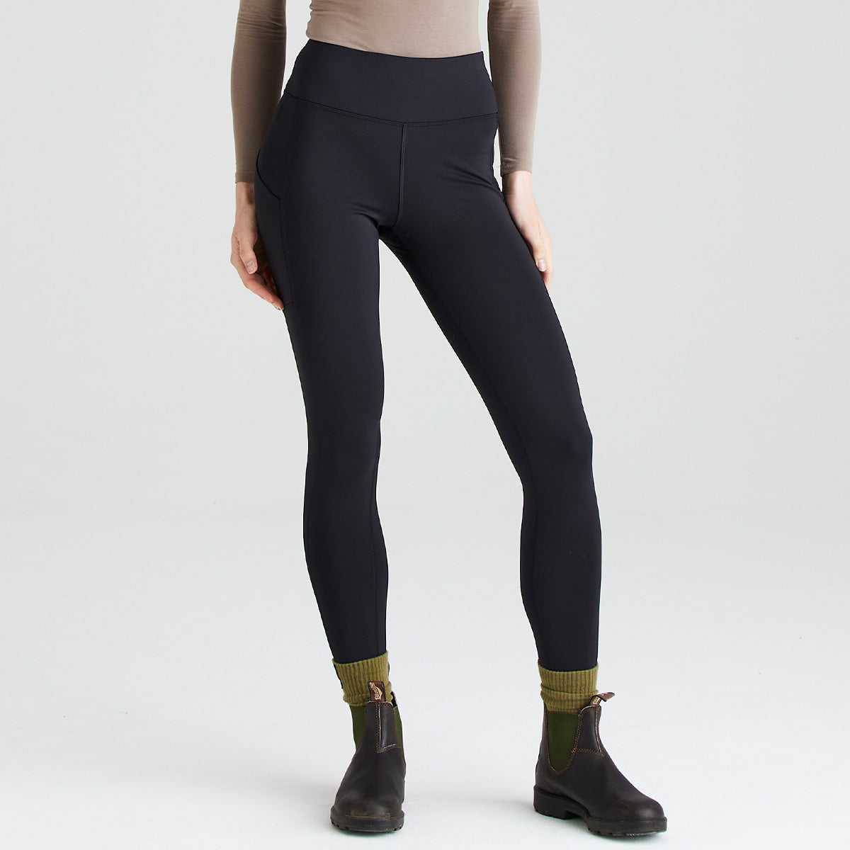 front view of a person wearing black high-waisted performance leggings paired with olive-green socks and dark ankle boots. The leggings feature a smooth, fitted design with a wide waistband and side pocket detail on the thigh. The image is photographed against a plain light gray studio background, highlighting the fit, stretch fabric, and streamlined silhouette of the leggings.
