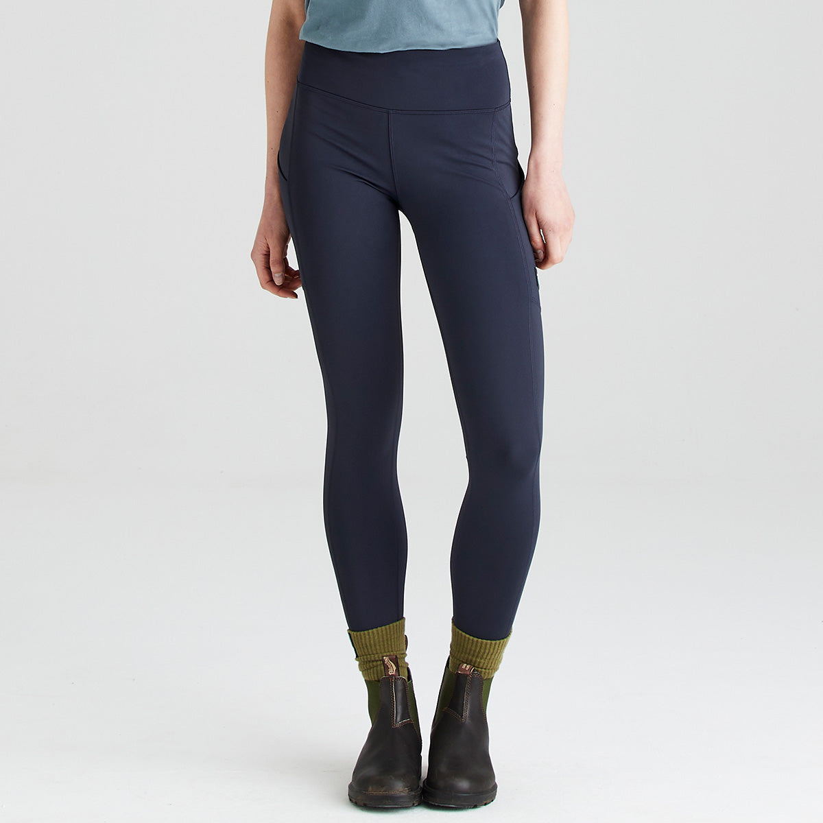 Front view of a person wearing Ridgeline navy high-waisted performance leggings with a wide supportive waistband and practical side thigh pockets. The leggings are paired with olive-green socks, dark ankle boots, and a blue short-sleeve top. Photographed against a plain light grey studio background, the image highlights the streamlined fit, stretch fabric, and functional design of the Ridgeline leggings for outdoor activities and everyday wear.
