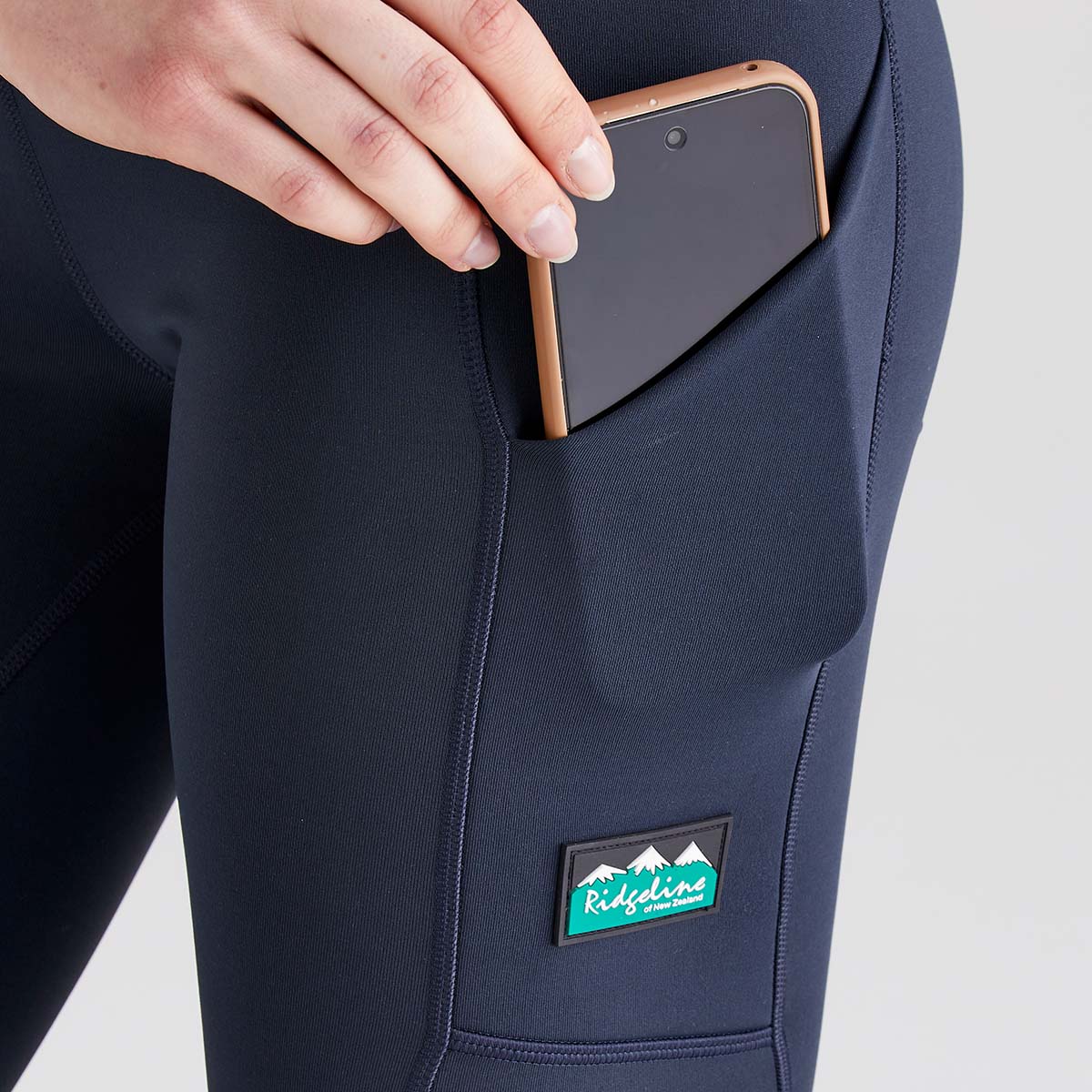 Close-up detail of Ridgeline navy performance leggings showing a side thigh pocket with a smartphone partially inserted for easy access. The leggings feature smooth stretch fabric, contoured seam detailing, and a fitted design for comfort and mobility. A Ridgeline New Zealand logo patch is positioned below the pocket, highlighting the practical storage feature and branded finish. Photographed against a plain light grey studio background.
