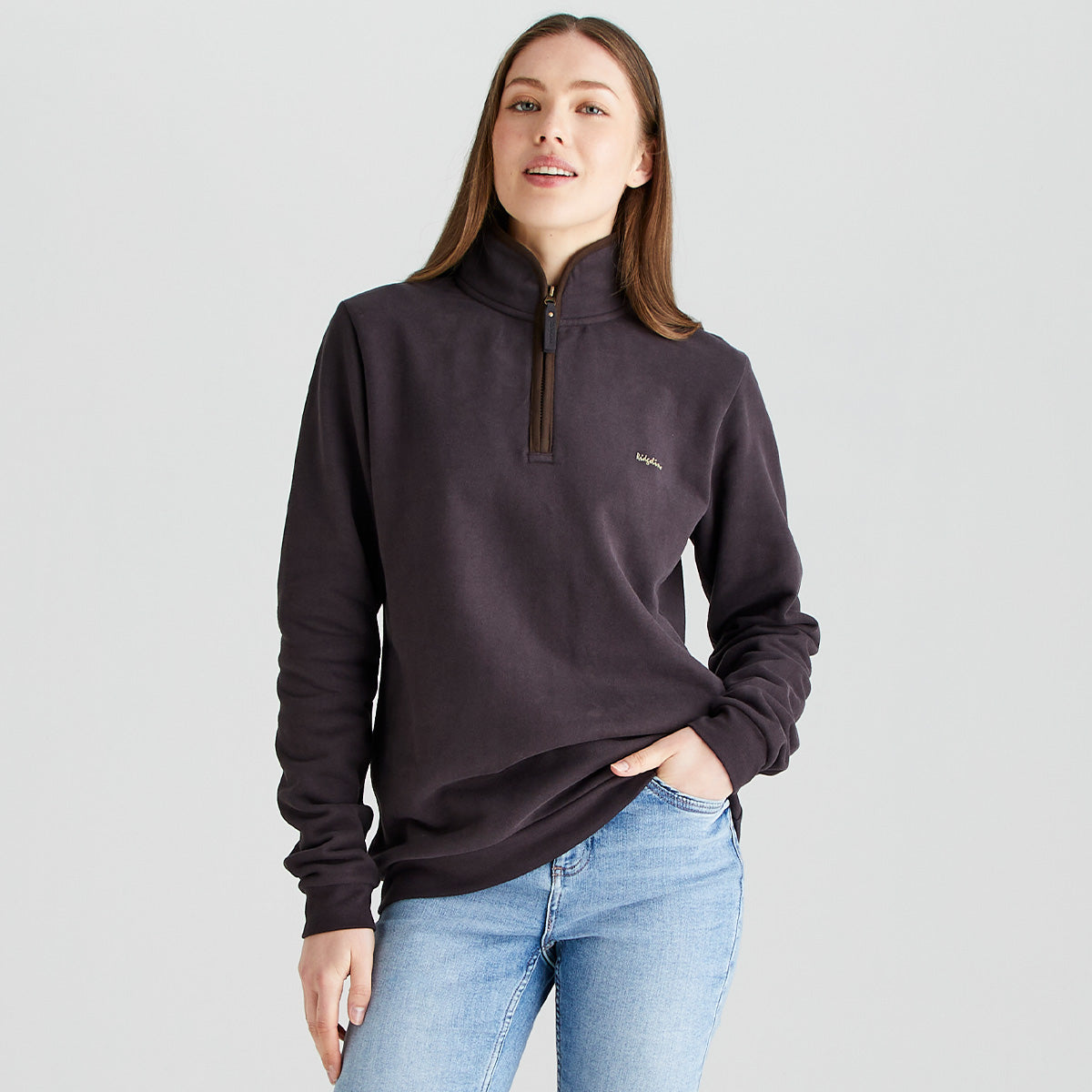 Person standing against a light gray studio background, wearing a dark brown quarter-zip sweatshirt with a high collar and light blue jeans. One hand is in a jeans pocket while the other rests by the side. The sweatshirt features a small embroidered logo on the left chest. The image is evenly lit and shows the outfit from about mid-thigh upward.
