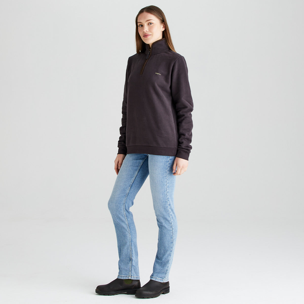 Full-length studio image of a person modeling a dark brown quarter-zip sweatshirt with a high collar, paired with light blue straight-leg jeans and black ankle boots. The person is standing at a slight angle against a light gray background, providing a three-quarter view of the sweatshirt's fit and silhouette. A small embroidered logo is visible on the left chest. The lighting is soft and even, highlighting the garment's color, fabric texture, and overall shape.
