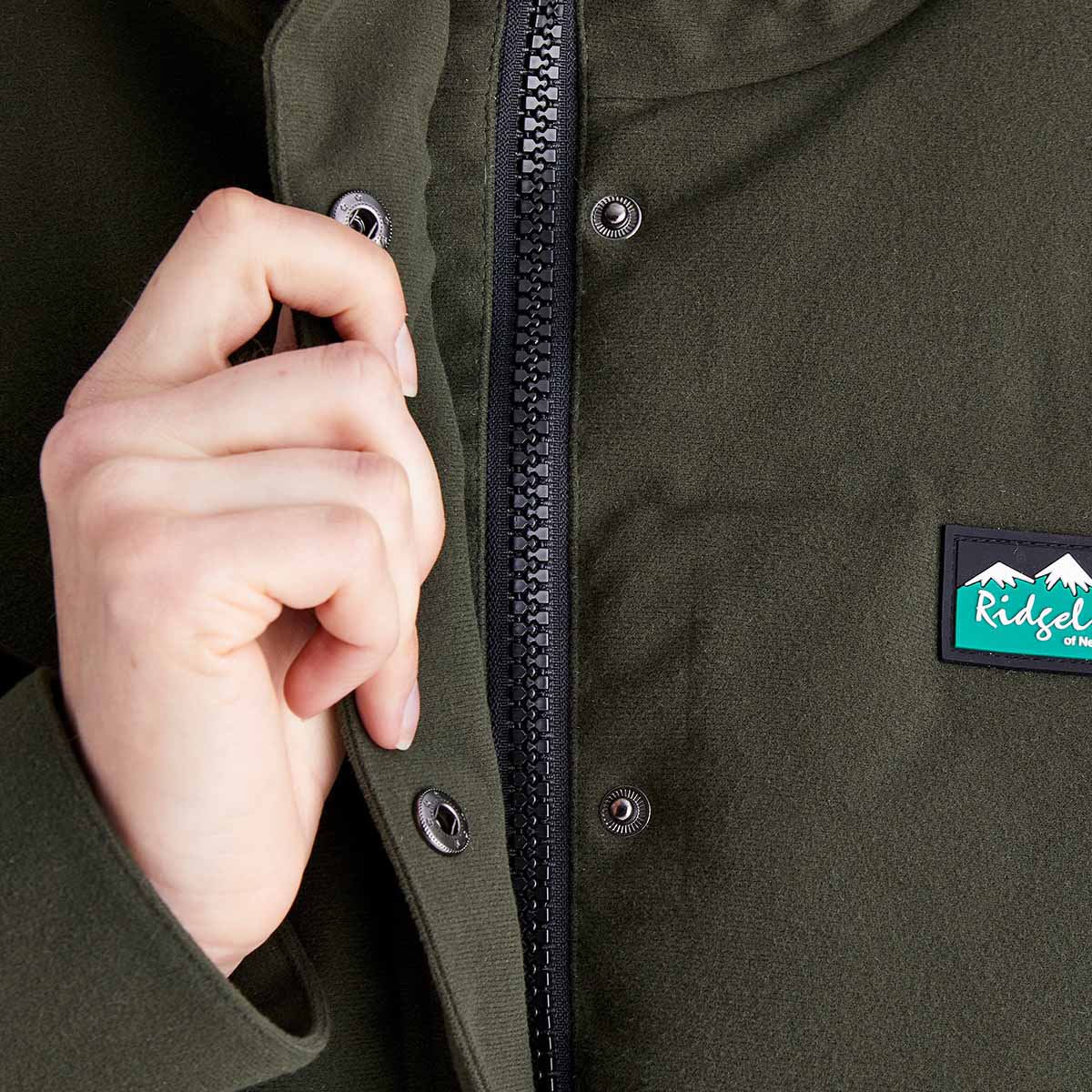 Close-up detail of a Ridgeline olive green insulated jacket showing the full-length front zip, snap-storm flap fastening and Ridgeline chest logo patch.
