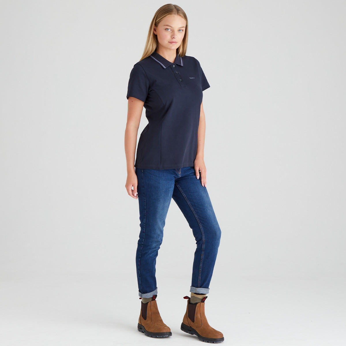 Woman wearing a navy polo shirt and blue jeans with brown boots on a light gray background