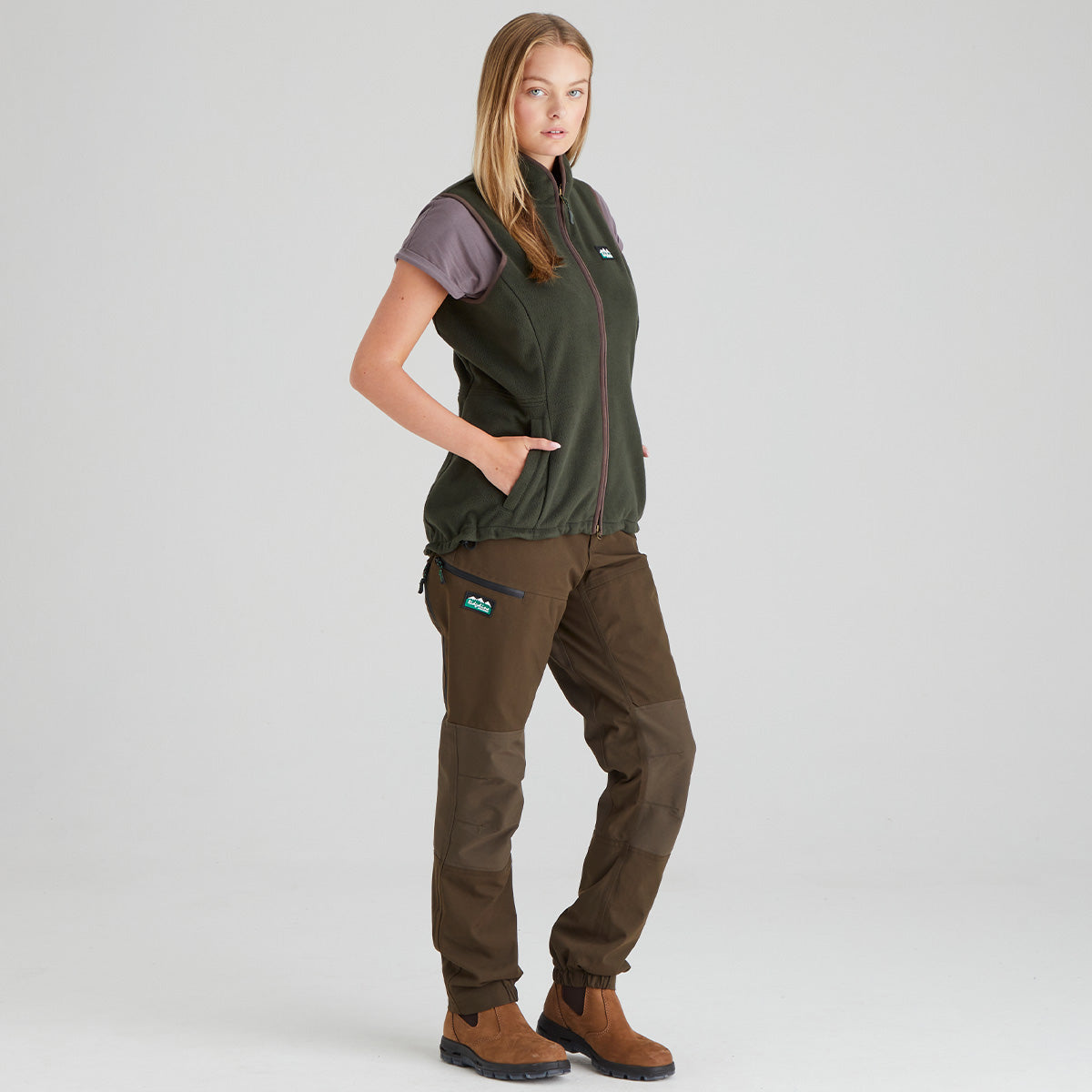 Woman wearing a green fleece gilet and brown pants on a grey background