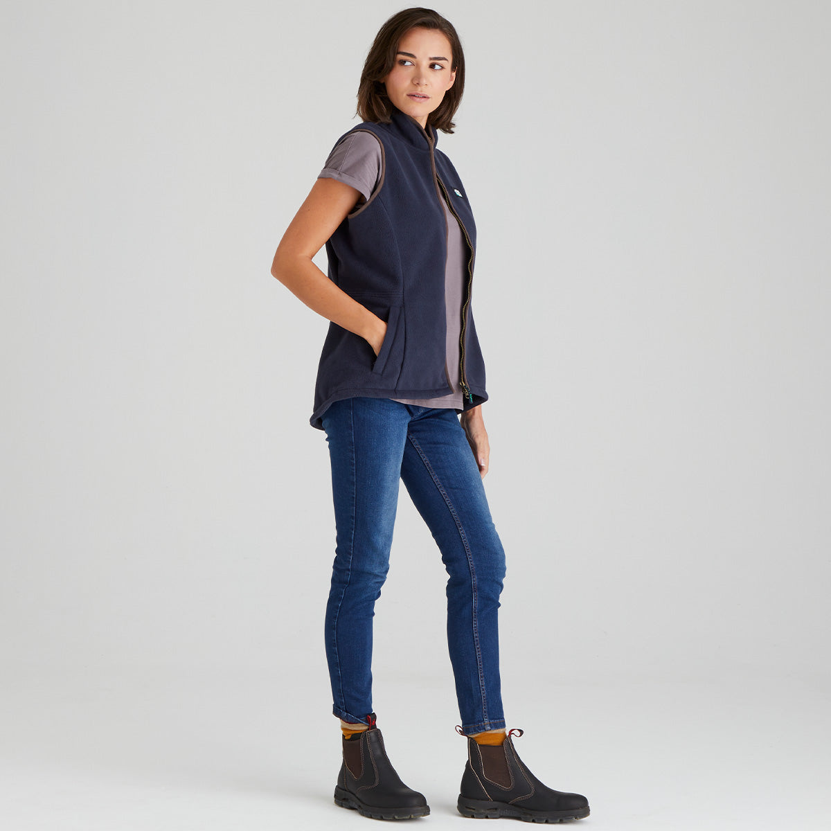 Woman wearing a navy vest, light shirt, blue jeans, and black boots on a white background