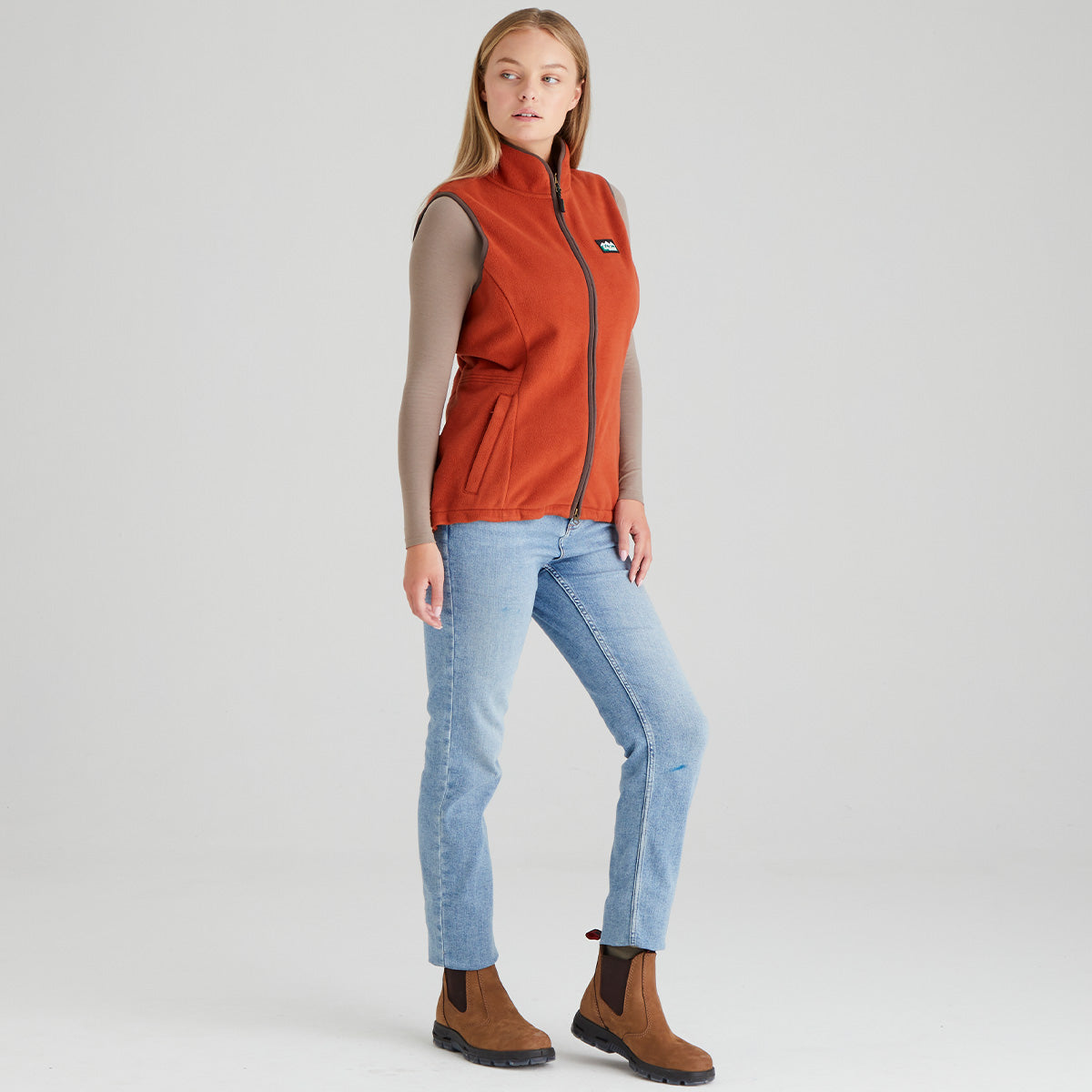 Woman wearing an orange vest, light blue jeans, and brown boots on a white background
