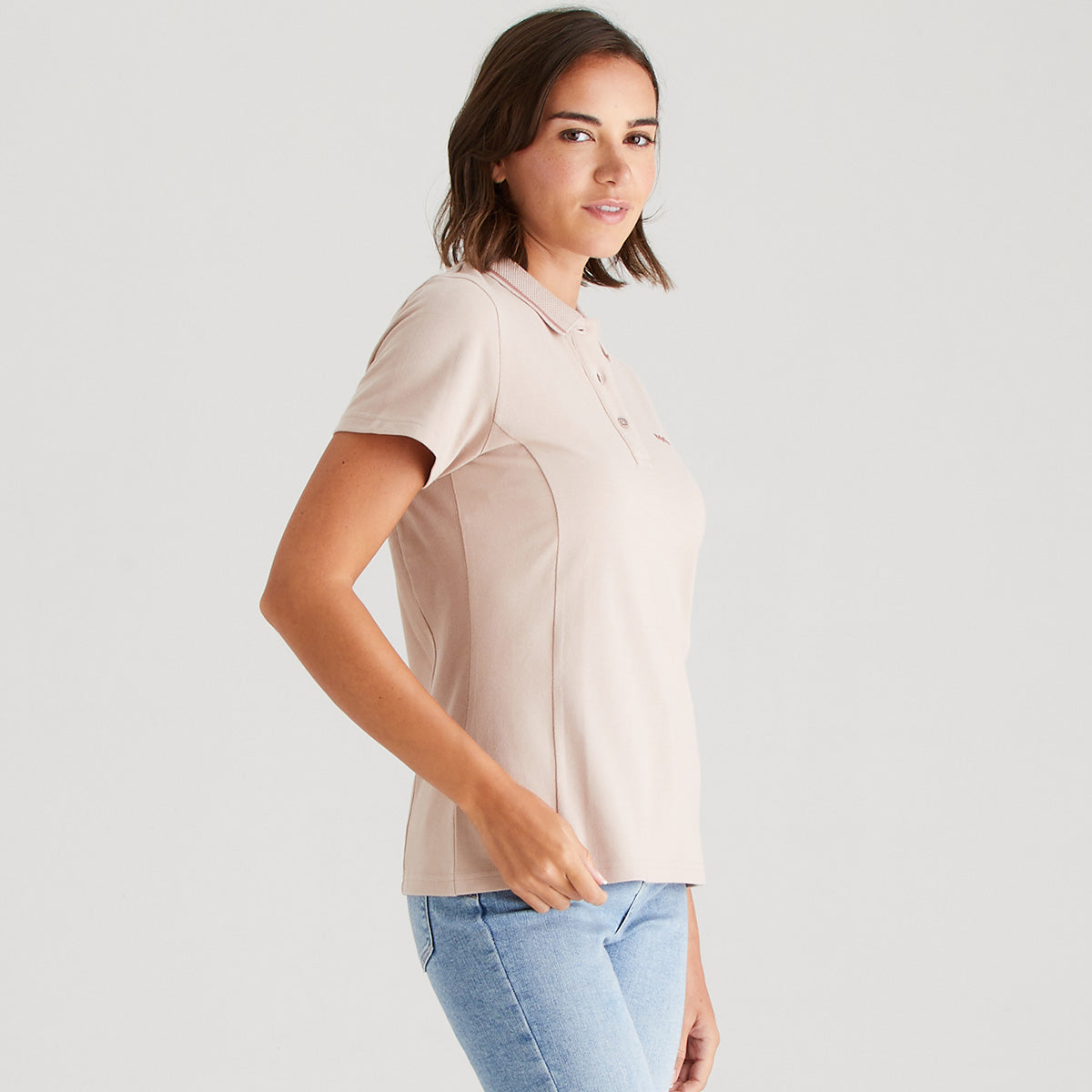 Woman wearing a blush polo shirt and light blue jeans on a plain background