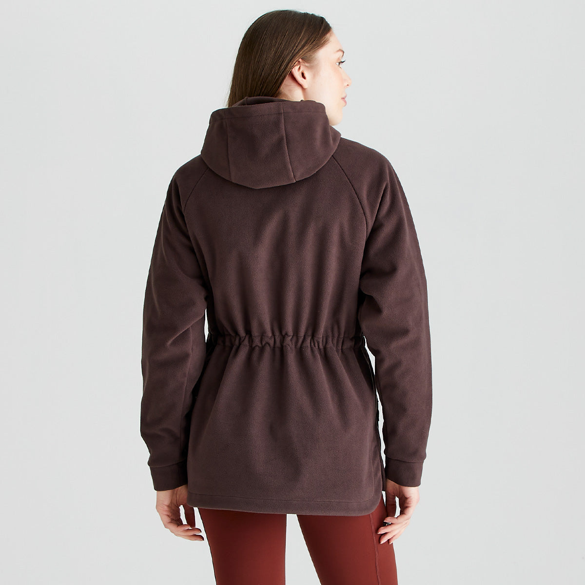 Rear view of a person wearing a Ridgeline dark brown or burgundy fleece smock with an attached hood, long raglan sleeves, and an adjustable drawcord waist. The soft fleece fabric and shaped silhouette are highlighted from the back, showcasing the gathered waist detail and extended hem coverage. Paired with rust-coloured performance leggings, the garment is photographed against a plain light grey studio background, emphasizing its comfortable fit and practical outdoor design.
