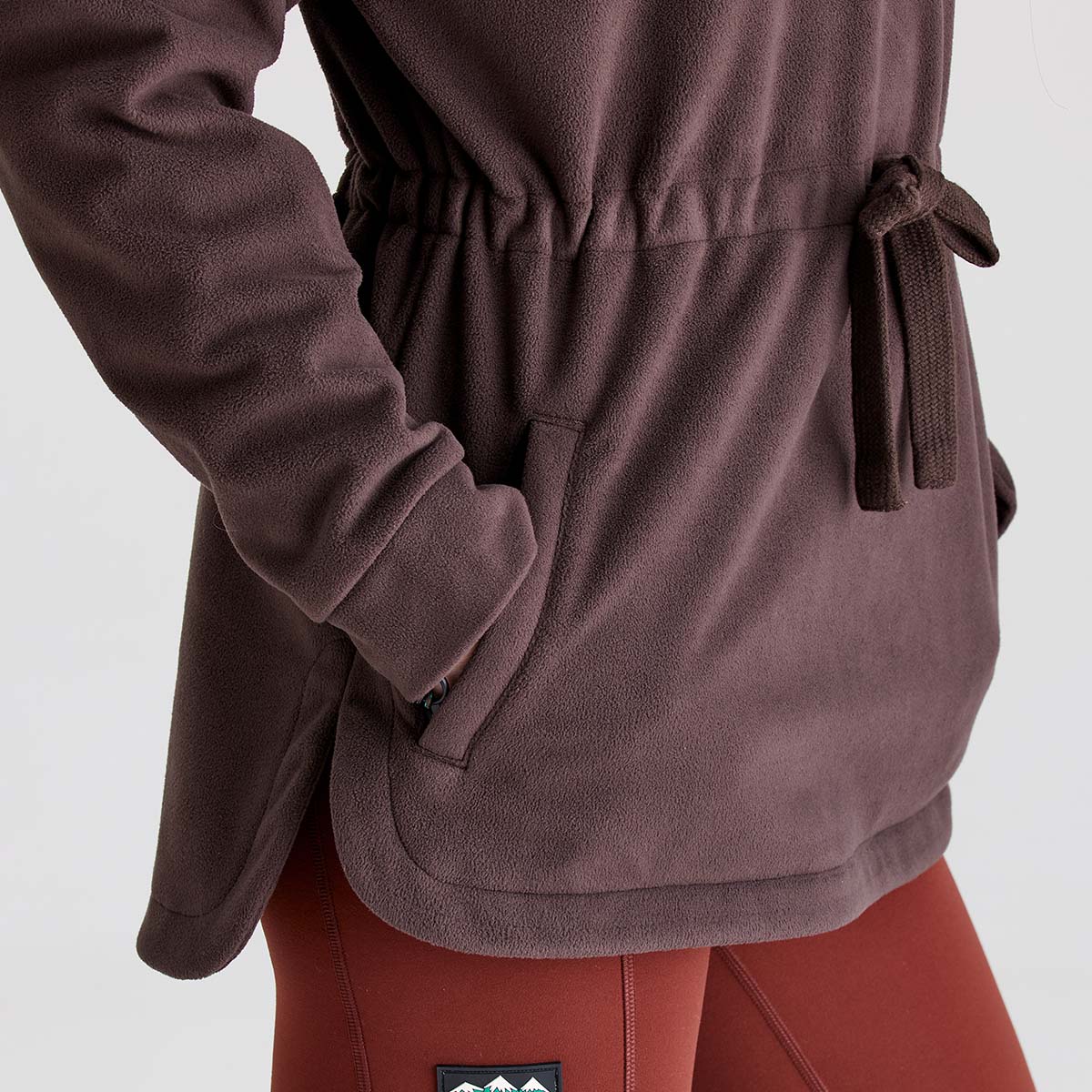 Close-up detail of a Ridgeline dark brown or burgundy fleece smock showing the adjustable drawcord waist, soft brushed fleece fabric, and hand-warmer pocket. The image highlights the fleece's textured finish, practical pocket design, curved hem, and tailored waist adjustment for a comfortable fit. Paired with rust-coloured leggings, the garment is photographed against a plain light grey studio background.
