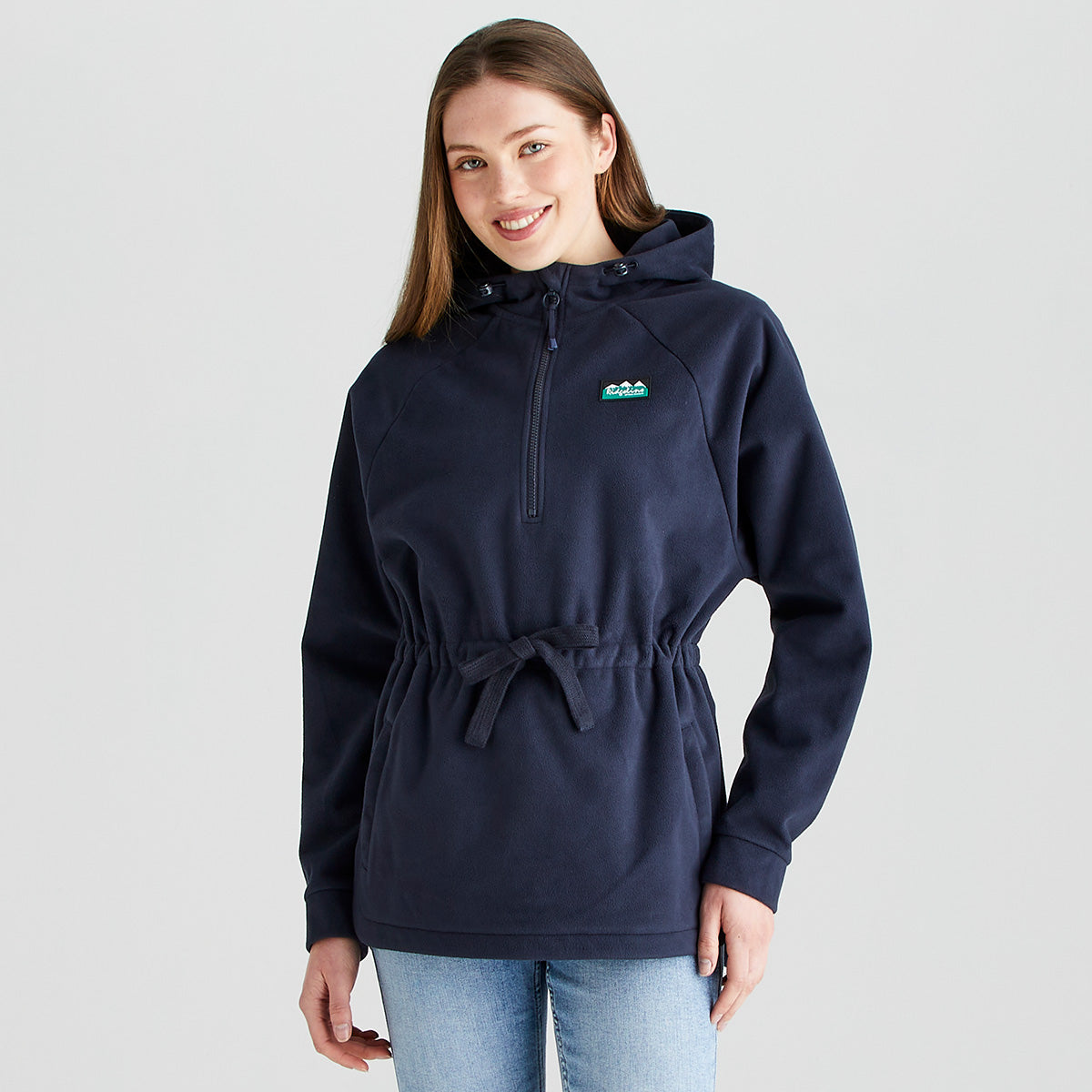 Front view of a person wearing a Ridgeline navy fleece smock with a half-zip neckline, adjustable drawcord waist, and attached hood. The fleece features a Ridgeline logo patch on the chest, long raglan sleeves, and a soft brushed finish for warmth and comfort. Paired with light blue jeans, the garment is photographed against a plain light grey studio background, highlighting the relaxed fit, practical outdoor styling, and adjustable waist detail.
