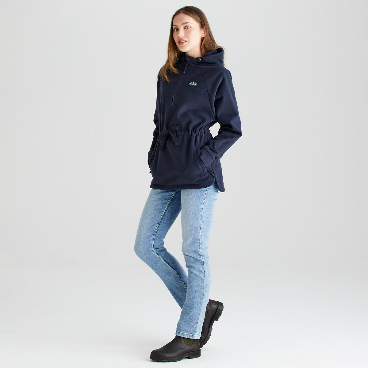 Full-length studio image of a person wearing a Ridgeline navy fleece smock with a half-zip neckline, attached hood, adjustable drawcord waist, and zipped hand pockets. The fleece is paired with light blue jeans and dark outdoor ankle boots. Photographed against a plain light grey background, the image highlights the smock's relaxed fit, curved hem, soft fleece fabric, and practical outdoor styling for everyday wear.
