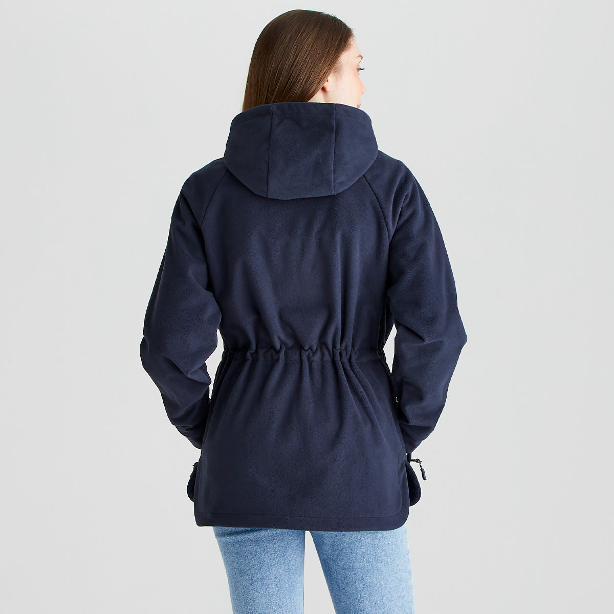 Rear view of a person wearing a Ridgeline navy fleece smock with an attached hood, long raglan sleeves, and an adjustable drawcord waist. The soft brushed fleece fabric and gathered waist detail create a shaped silhouette, while zipped hand pockets are visible at the sides. Paired with light blue jeans, the garment is photographed against a plain light grey studio background, highlighting the back fit, hood design, and practical outdoor styling.

