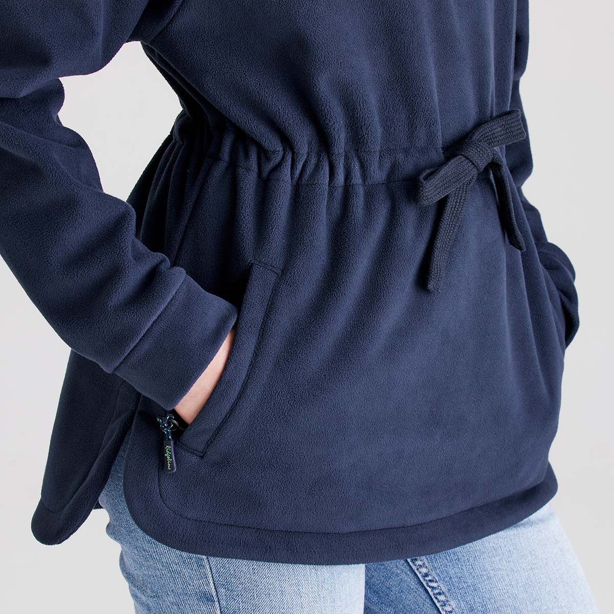 Close-up detail of a Ridgeline navy fleece smock showing the adjustable drawcord waist, zipped hand-warmer pocket, and soft brushed fleece fabric. The image highlights the gathered waist design, practical pocket construction, curved hem, and warm fleece texture. Photographed against a plain light grey studio background, the close-up showcases the smock's functional outdoor features and comfortable fit.
