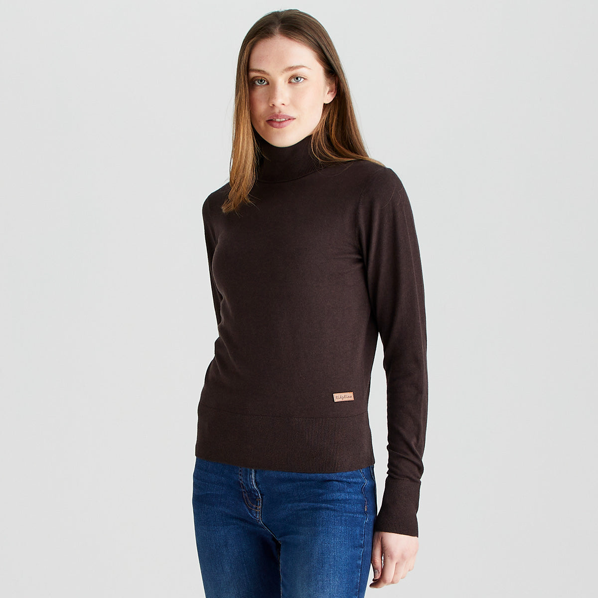 Woman modelling a Ridgeline knitted roll neck jumper in dark brown, featuring a high folded turtleneck, long sleeves, ribbed cuffs and hem, and a fitted silhouette. Shown against a light grey background with blue jeans.
