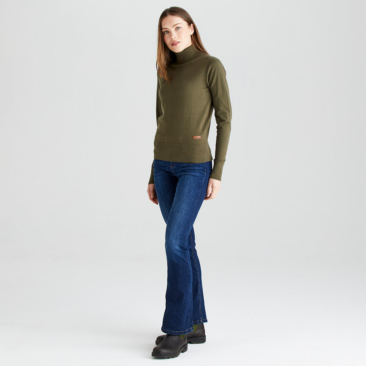 Full-length angled view of a model wearing a Ridgeline knitted roll neck pullover in olive green. The fine-knit jumper features a high roll-neck collar, long sleeves, and a ribbed hem and cuffs, finished with a small Ridgeline logo tag near the waist. Styled with blue jeans and black ankle boots against a light grey studio background.