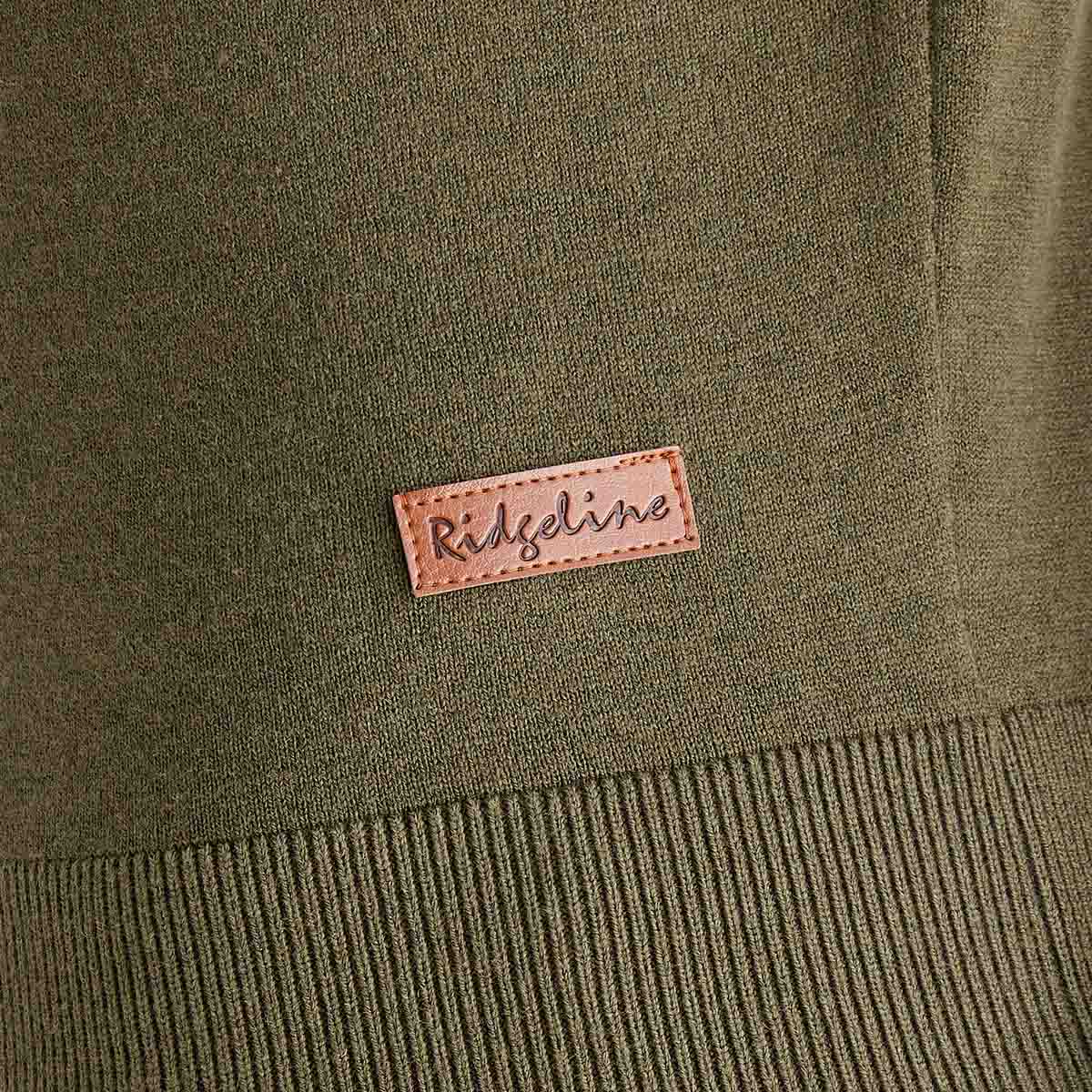Close-up detail of an olive green Ridgeline knitted roll neck pullover, highlighting the fine-knit fabric, ribbed hem, and stitched tan leather Ridgeline logo patch near the lower front. The image showcases the jumper's texture and premium finishing.
