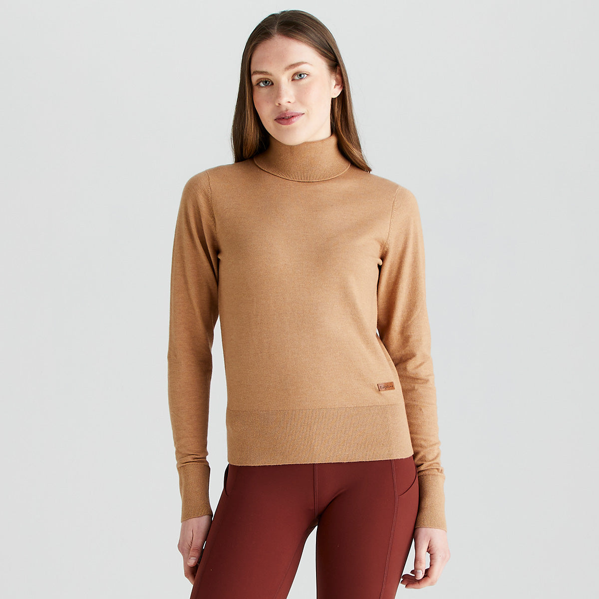 Front view of a model wearing a Ridgeline knitted roll neck pullover in camel brown. The fine-knit jumper features a high roll-neck collar, long sleeves, and a ribbed hem and cuffs, with a small Ridgeline logo tag near the waist. Styled with burgundy leggings against a light grey studio background.
