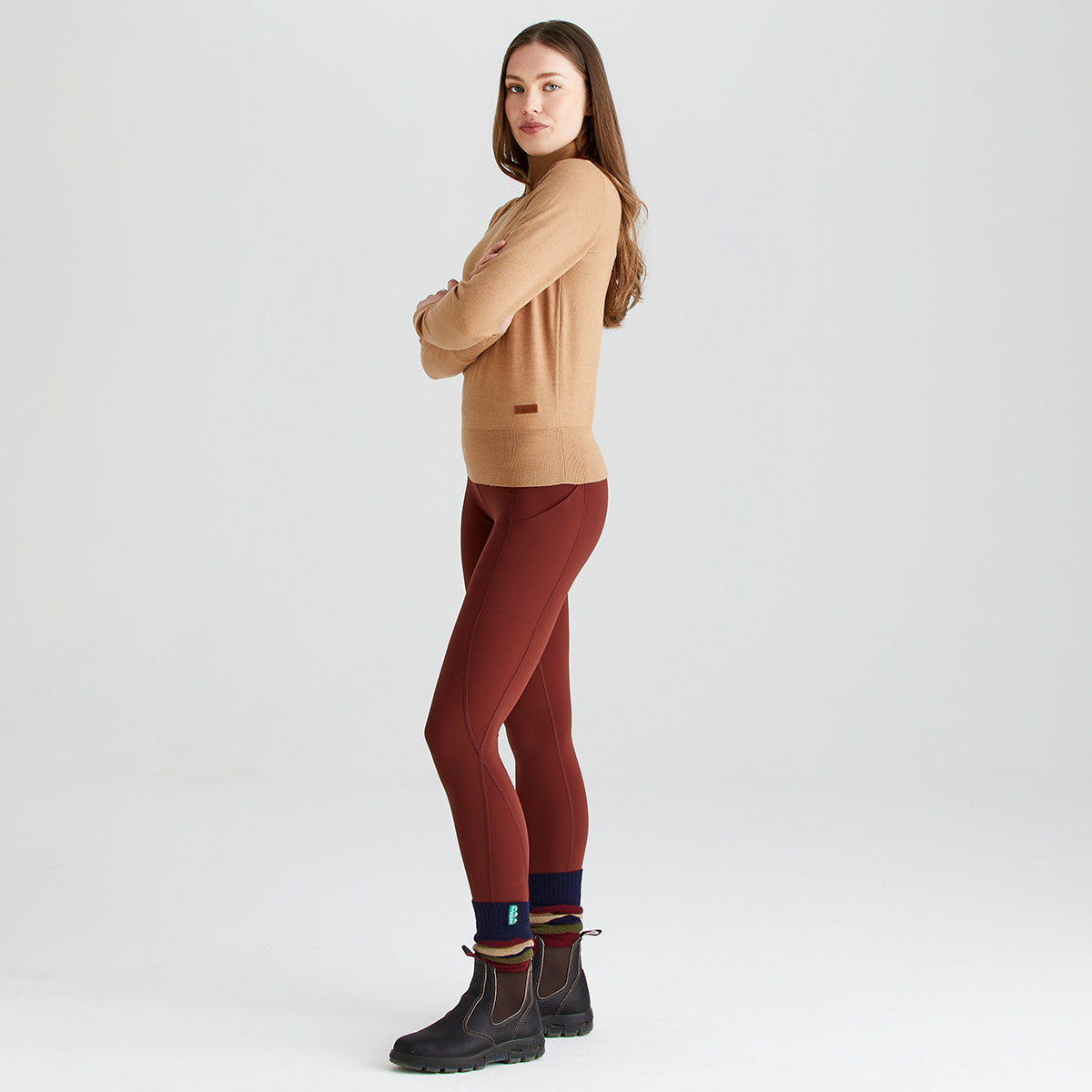 Side-profile full-length view of a model wearing a Ridgeline knitted roll neck pullover in camel brown. The fine-knit jumper features a high roll-neck collar, long sleeves, ribbed hem and cuffs, and a small Ridgeline logo tag near the waist. Styled with burgundy leggings, patterned socks, and brown ankle boots against a light grey studio background.
