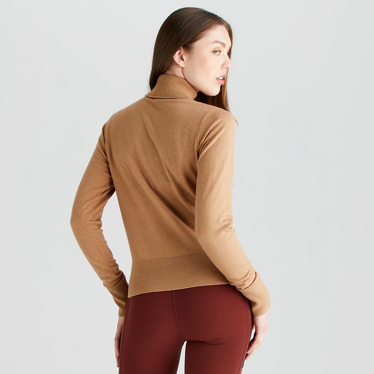 Rear angled view of a model wearing a Ridgeline knitted roll neck pullover in camel brown. The fine-knit jumper features a high roll-neck collar, long sleeves, and a wide ribbed hem for a comfortable regular fit. Styled with burgundy leggings against a light grey studio background, highlighting the back fit and silhouette of the garment.

