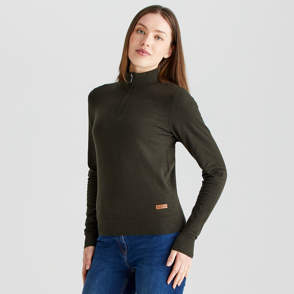 A person modeling a dark olive-green long-sleeve knit top with a quarter-zip high collar, paired with blue jeans. The person is standing against a plain light gray studio background, with one hand resting at the side and the other near the hip. The fitted sweater features ribbed cuffs and hem and a small brand label near the lower hem. The image is evenly lit, highlighting the texture and shape of the garment.
