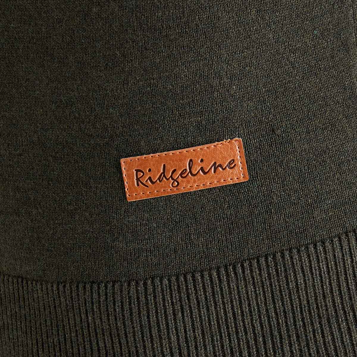 Close-up detail of a dark olive-green knit sweater showing the ribbed hem and a small rectangular brown faux-leather brand patch stitched near the bottom. The patch is embossed with the text “Ridgeline.” The image highlights the fabric texture, stitching, and garment branding against the smooth knit material.
