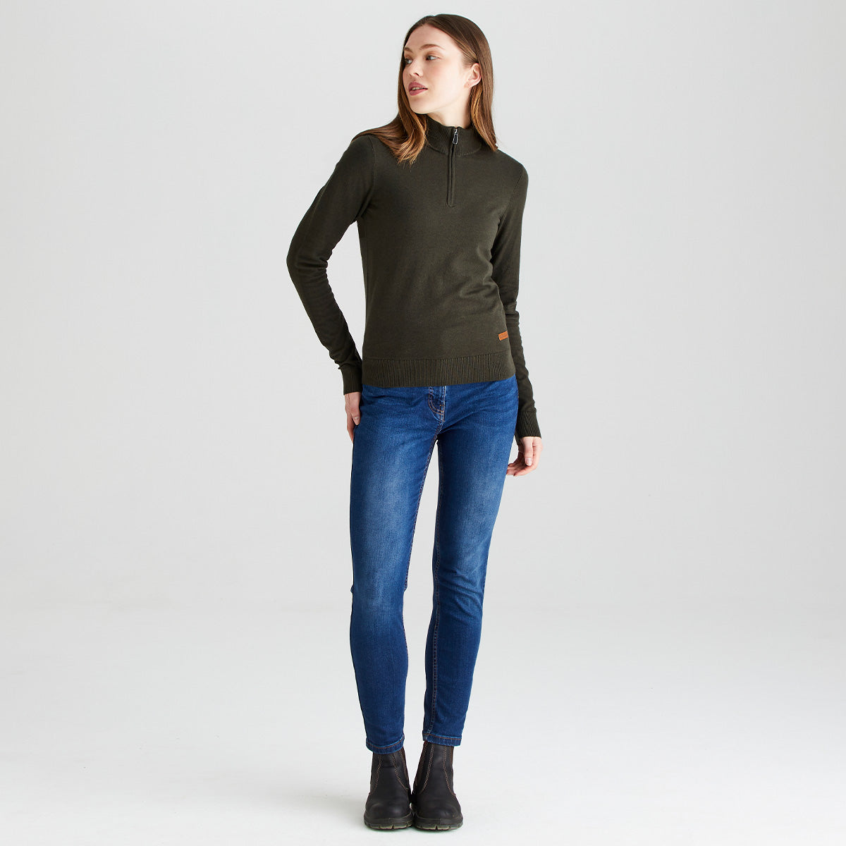 Full-length studio image of a person wearing a dark olive-green quarter-zip knit sweater, fitted blue jeans, and dark ankle boots. The sweater features a high collar with a short front zip, long sleeves, and a ribbed hem. The person is standing against a plain light gray background in a relaxed pose. A small brown brand patch is visible near the lower hem of the sweater. At the bottom of the screenshot, a website editing overlay partially shows the text “Click image to set focal point.
