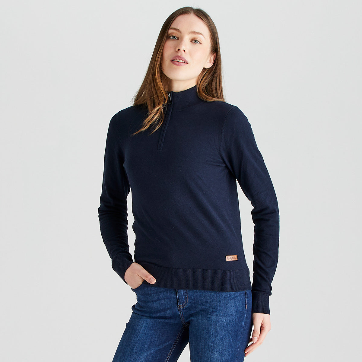 Person wearing a navy blue quarter-zip knit sweater with a high collar and long sleeves, paired with blue jeans. The sweater features a fine-knit construction, ribbed hem and cuffs, and a small branded patch near the lower hem. The person is standing against a plain light gray studio background with one hand in a pocket, highlighting the fit and style of the garment.
