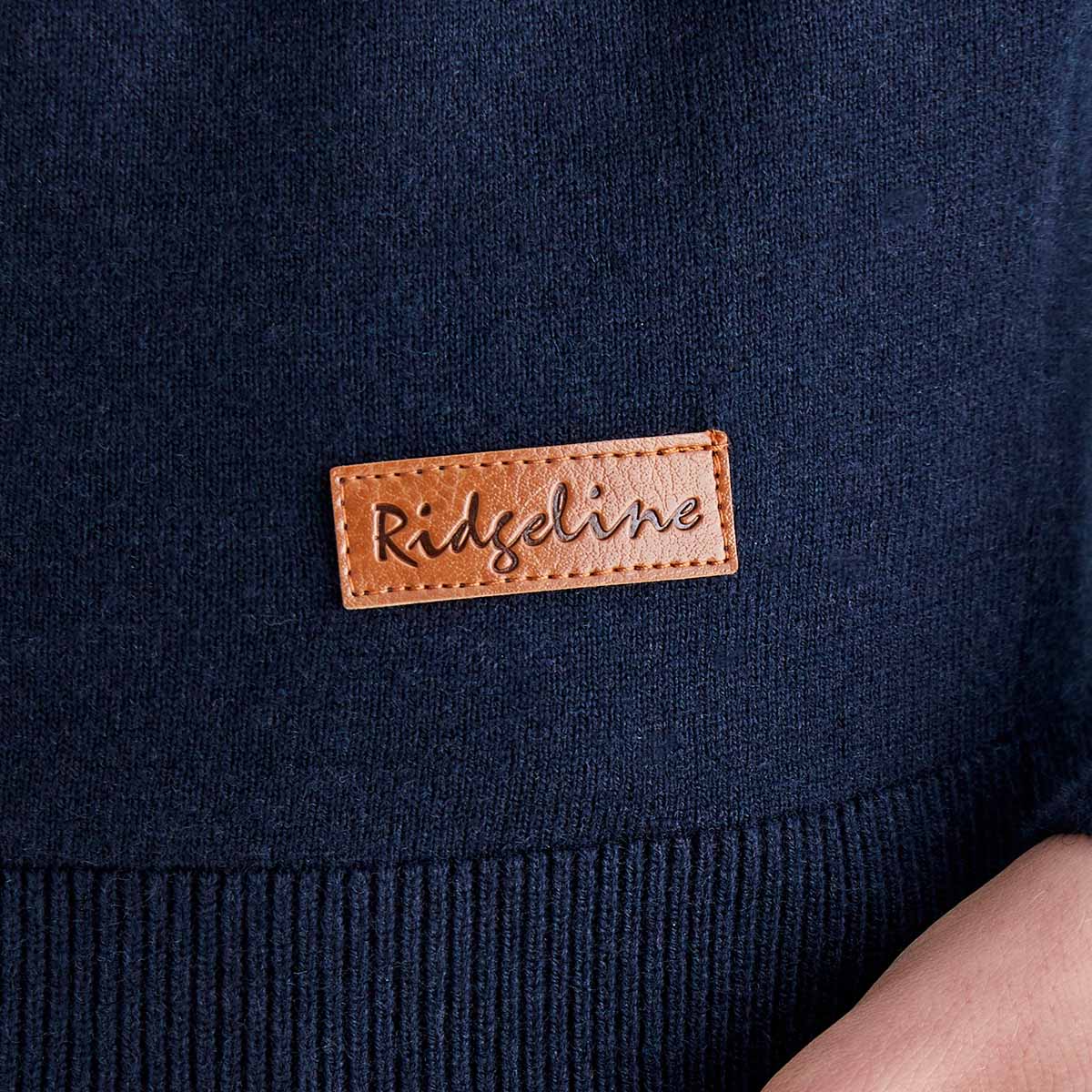 Close-up detail of a navy blue fine-knit sweater showing the ribbed hem and a brown faux-leather Ridgeline logo patch stitched near the lower hem. The image highlights the knit fabric texture, stitching, and branded label. Part of a hand is visible in the lower-right corner. 