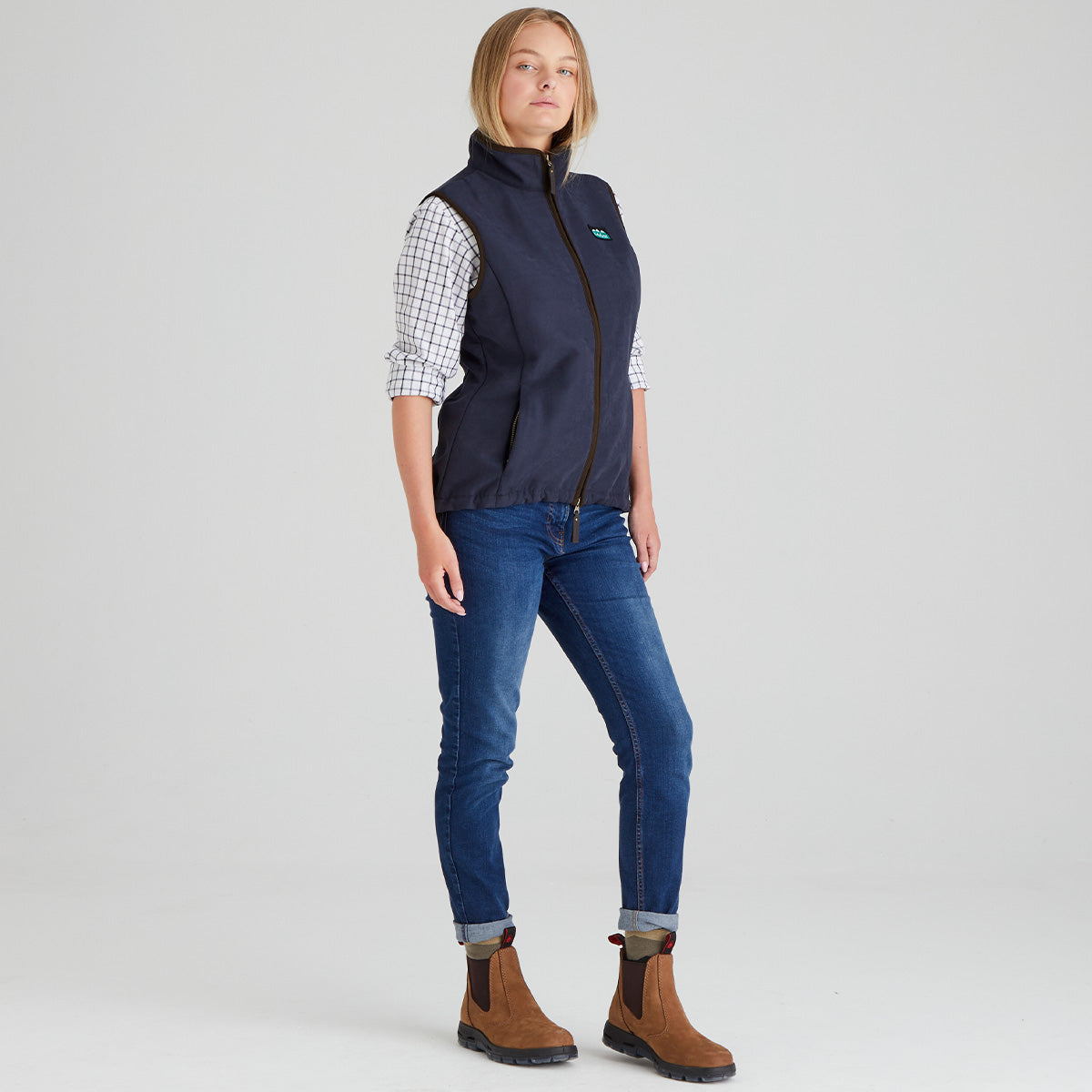 Person wearing a navy blue moleskin gilet vest over a plaid shirt, paired with blue jeans and brown boots on a light grey background.