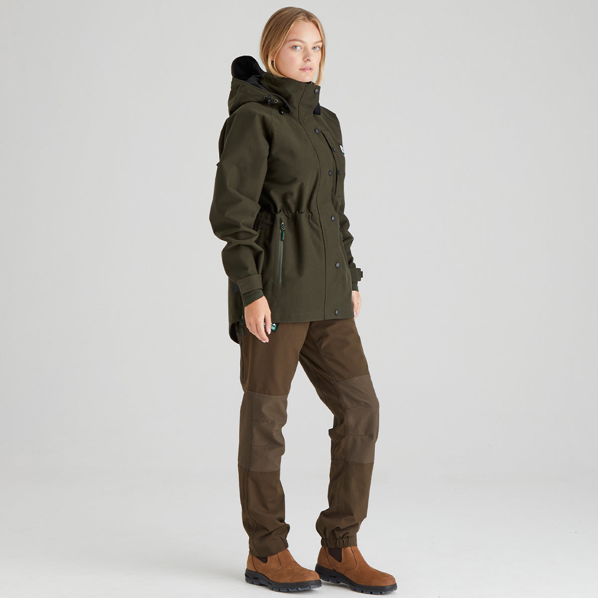 Woman wearing a green waterproof jacket and brown pants on a light grey background